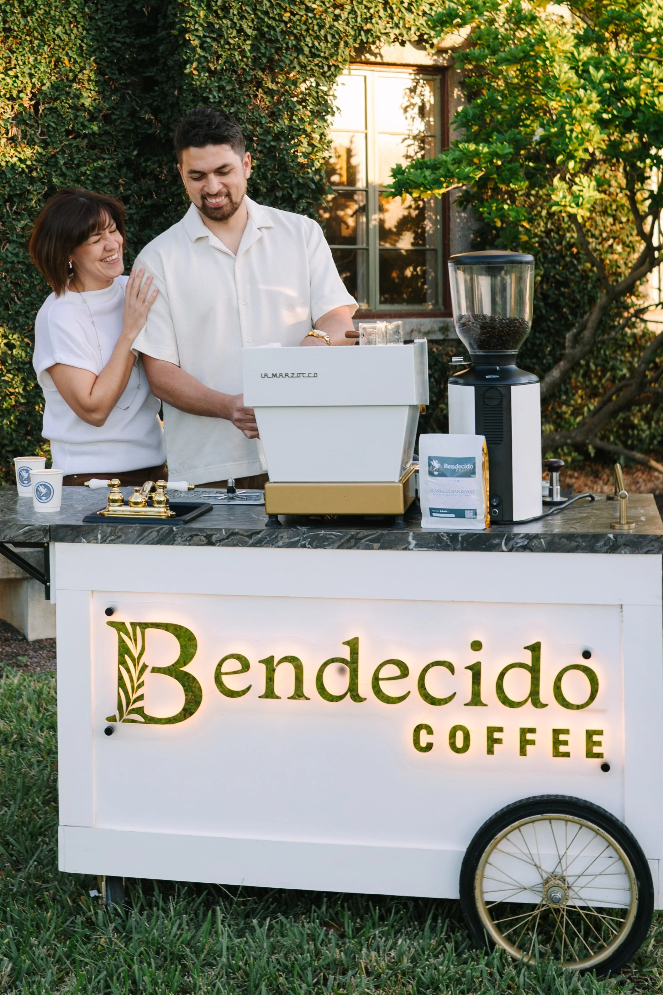 Two people, a man and a woman, enjoy making coffee at an outdoor coffee cart with a sign that reads "Bendecido Coffee". The woman is touching the man's shoulder, and they are smiling at each other. The coffee cart has a grinder with coffee beans, a c