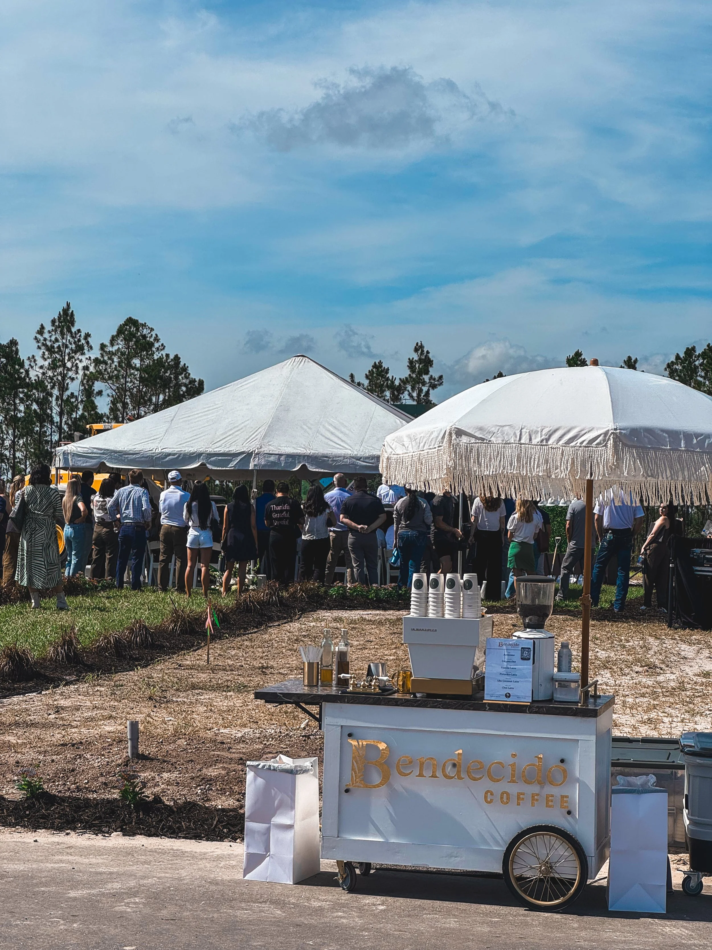 A coffee stand labeled 'Bendecido Coffee' with cups, bottles, and equipment on top, set outdoors on a sandy area under a white umbrella, with a large crowd gathered under tents in the background on a partly cloudy day.