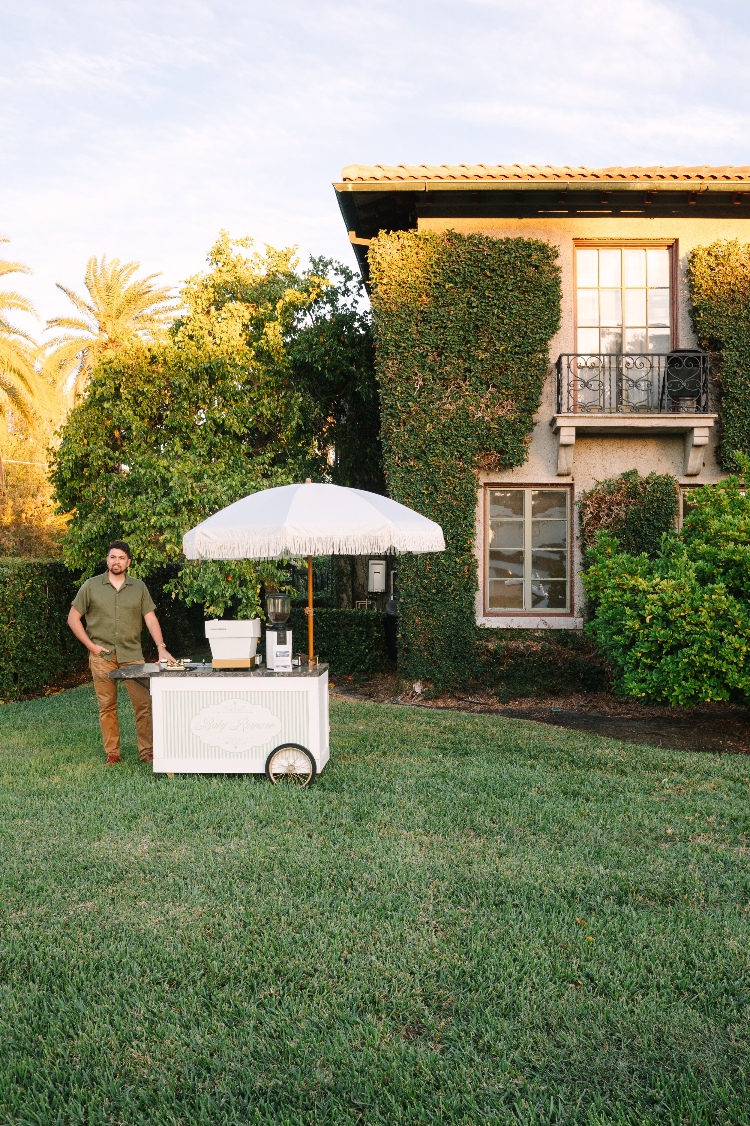 A man standing next to a white mobile coffee cart with an umbrella in a garden, with a tall house covered in green vines in the background, during late afternoon or early evening.