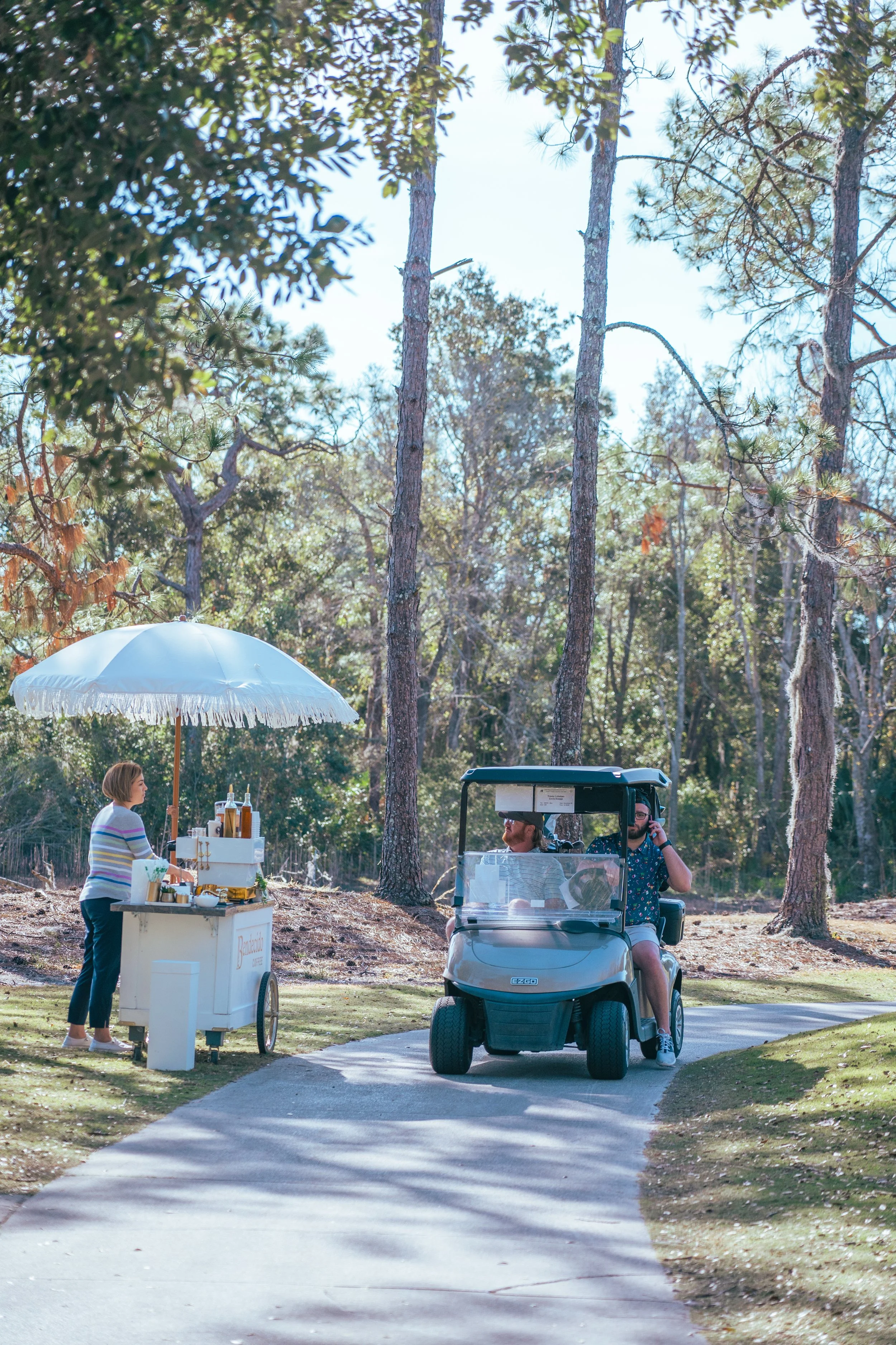 A person stands at a small, white mobile beverage cart with a white umbrella, serving drinks to two people in a golf cart on a winding pathway in a wooded outdoor setting.