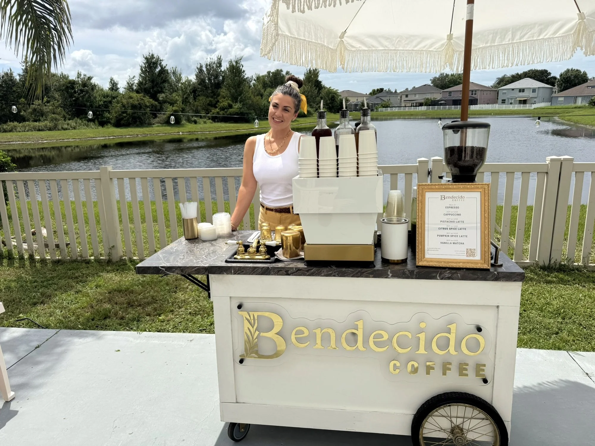 Barista at the Bendecido Coffee cart under a white umbrella at a lakeside outdoor event in Tampa Bay