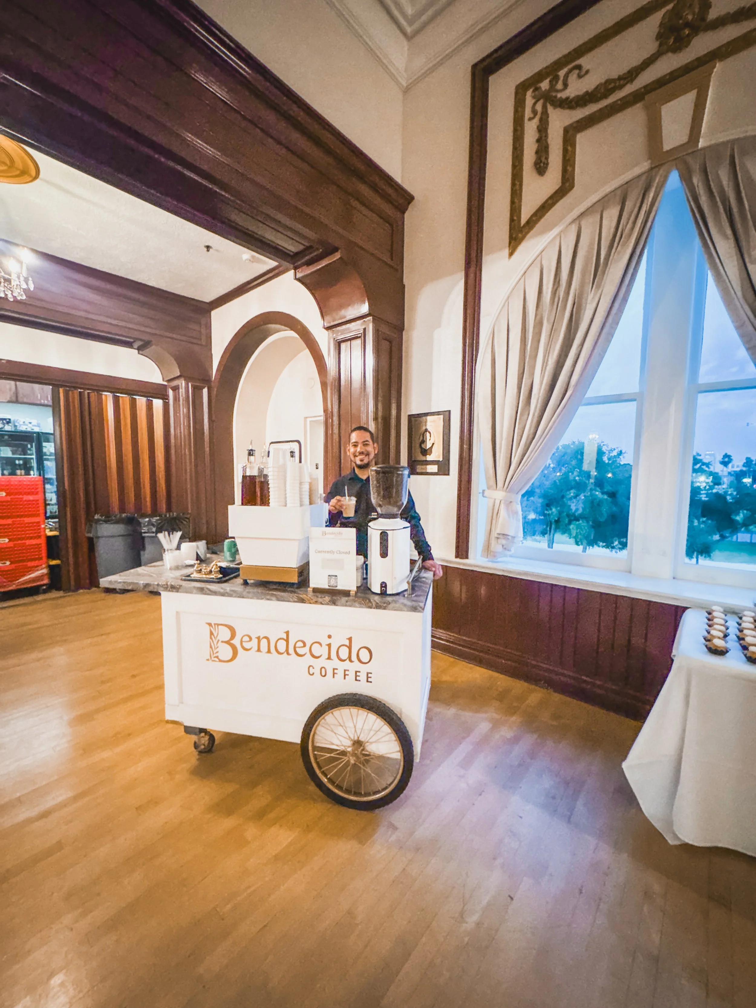 A smiling man standing behind a mobile coffee cart with ''Bendecido Coffee'' written on it, inside a room with large windows, wooden paneling, and beige walls.