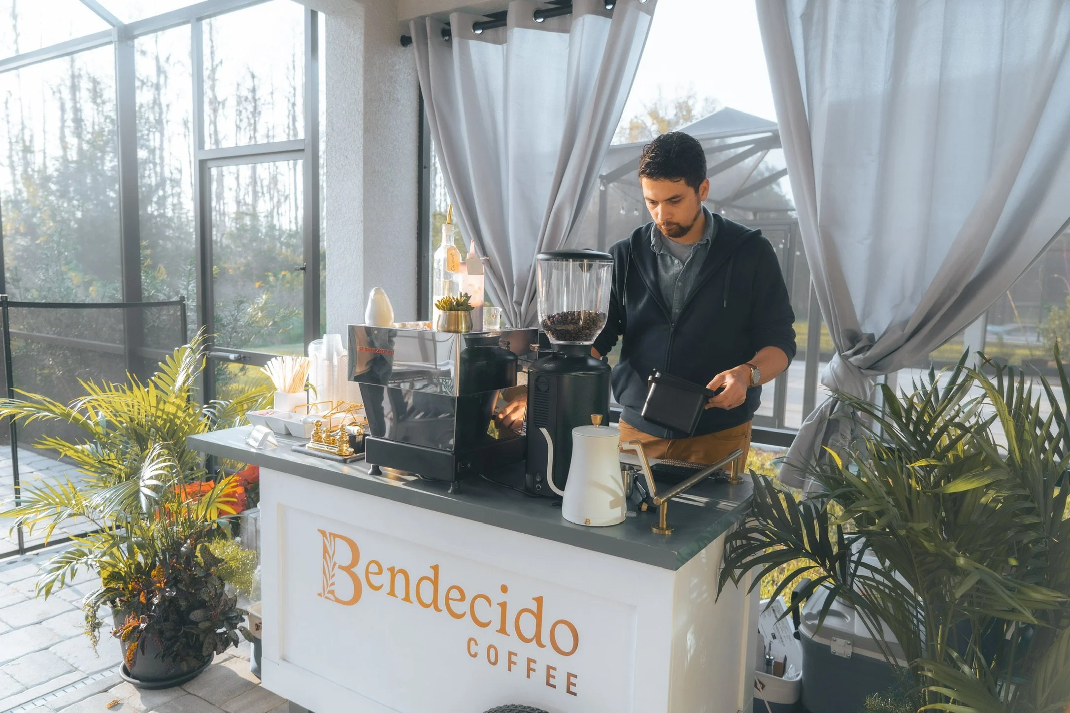 Barista working behind a Bendecido Coffee cart in a screened outdoor room with plants