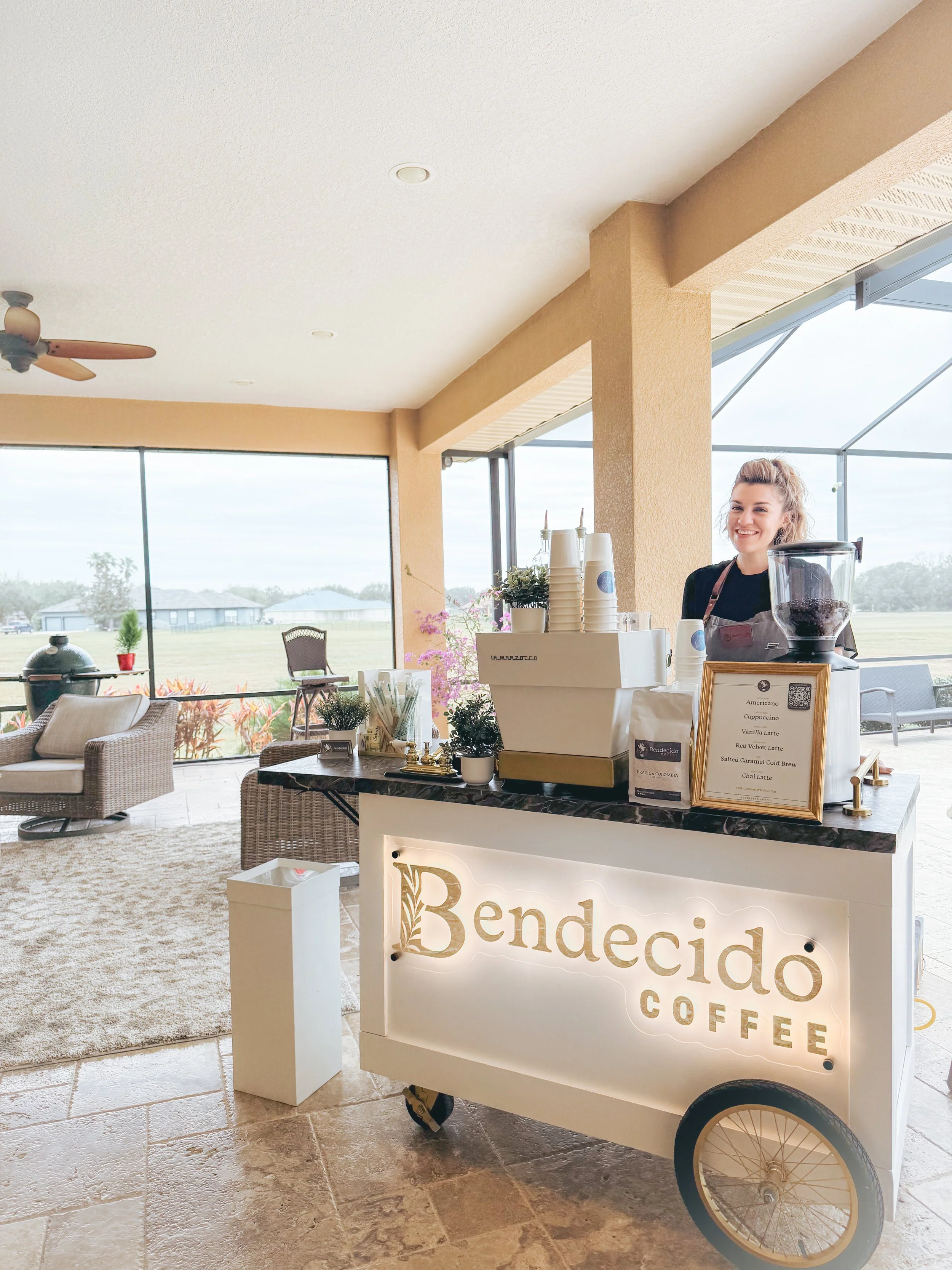 Smiling barista behind a white coffee cart with a sign that reads 'Bende cido Coffee,' inside a bright cafe with large windows, wicker chairs, and a view of a grassy field.