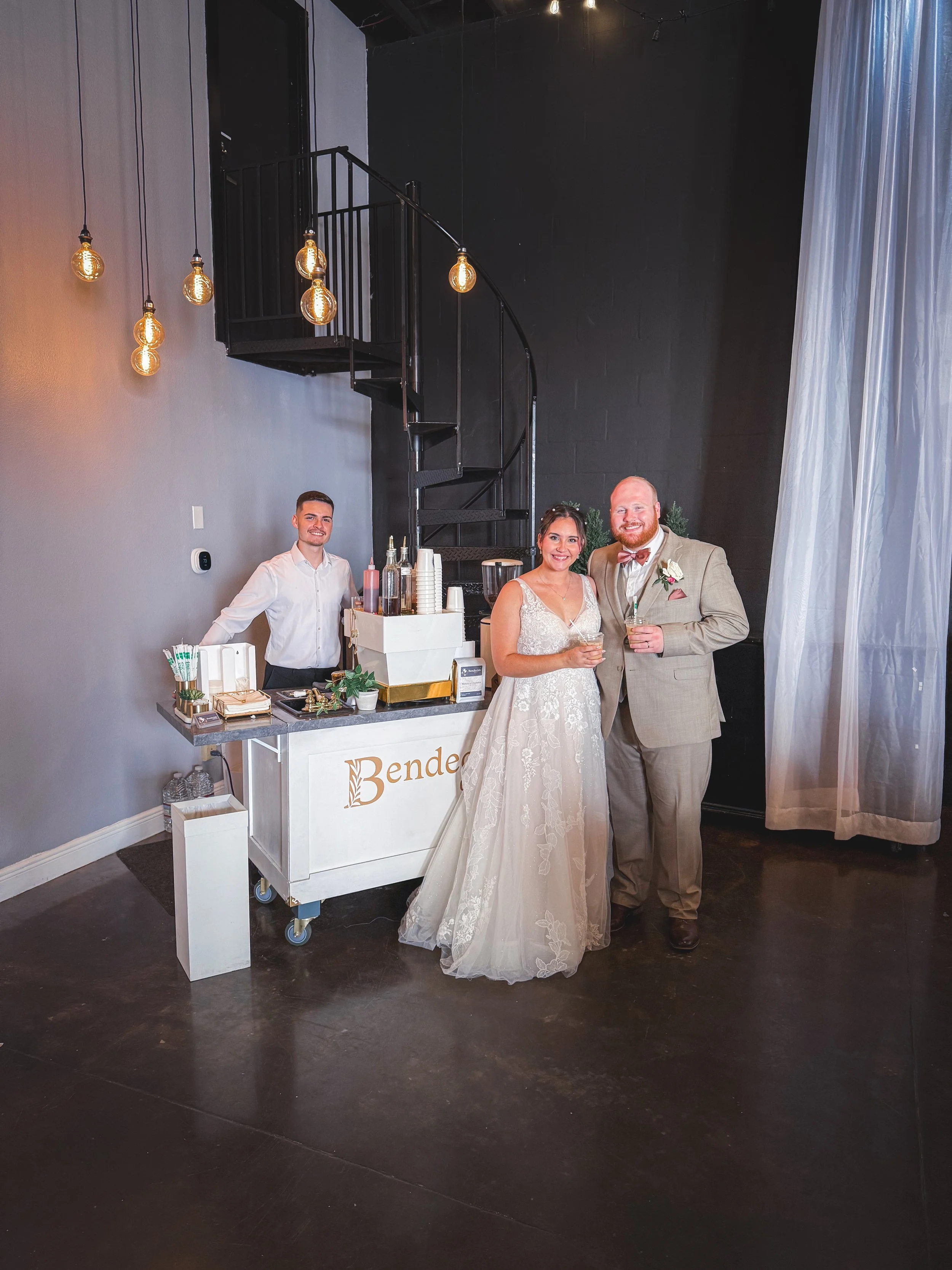 A bride and groom standing together at their wedding reception, holding drinks and smiling. They are positioned next to a white bar cart with the word "Bend" visible on it, and a bartender behind the cart. The scene features modern decor with hanging