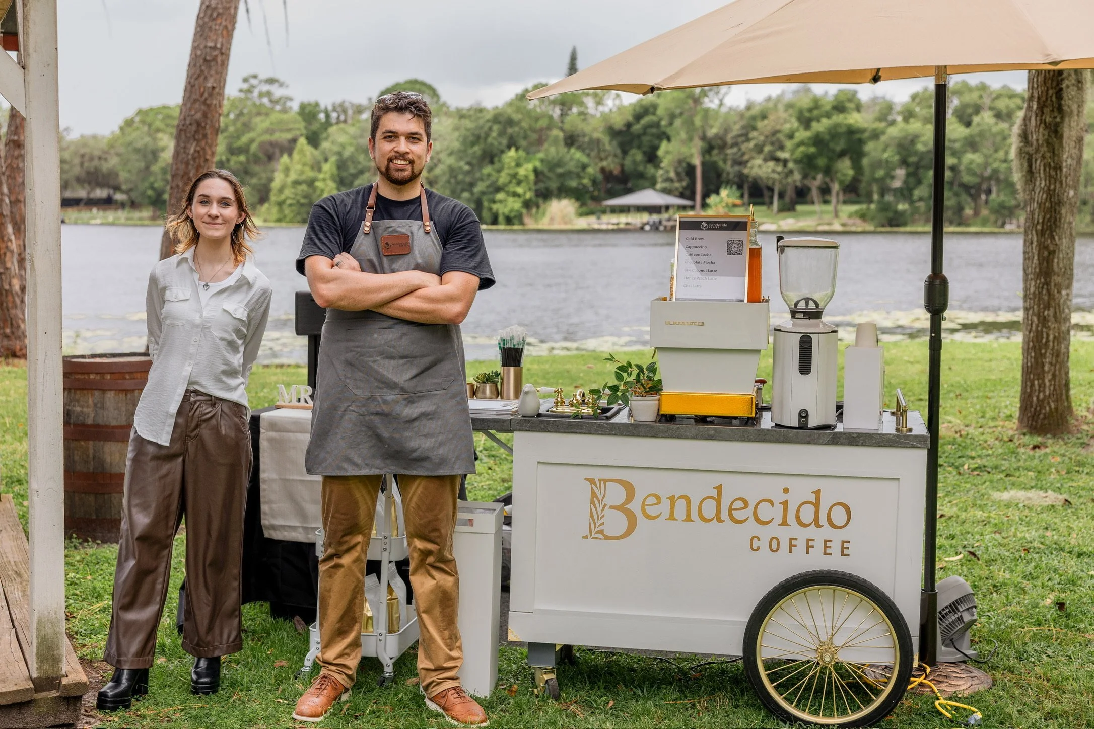 Group of people posing beside a Bendecido Coffee cart in a park by a lake