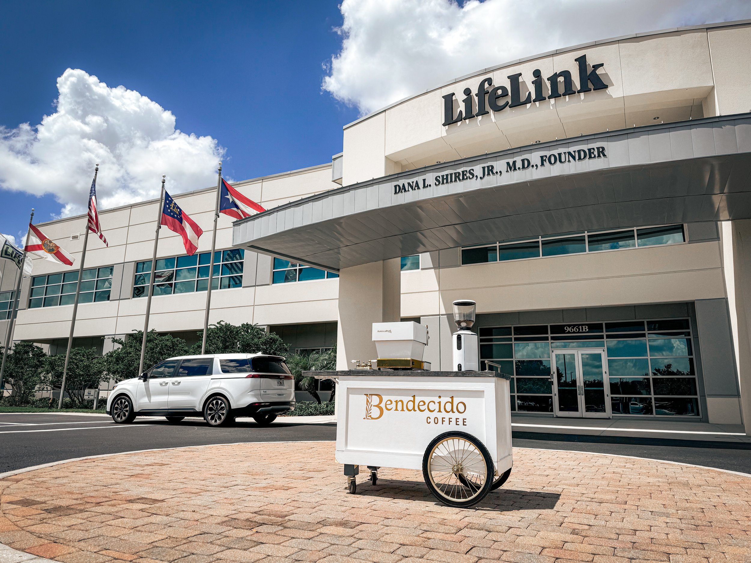 Exterior of LifeLink building with flags, a parked car, and a mobile coffee cart labeled 'Bendecido Coffee.'