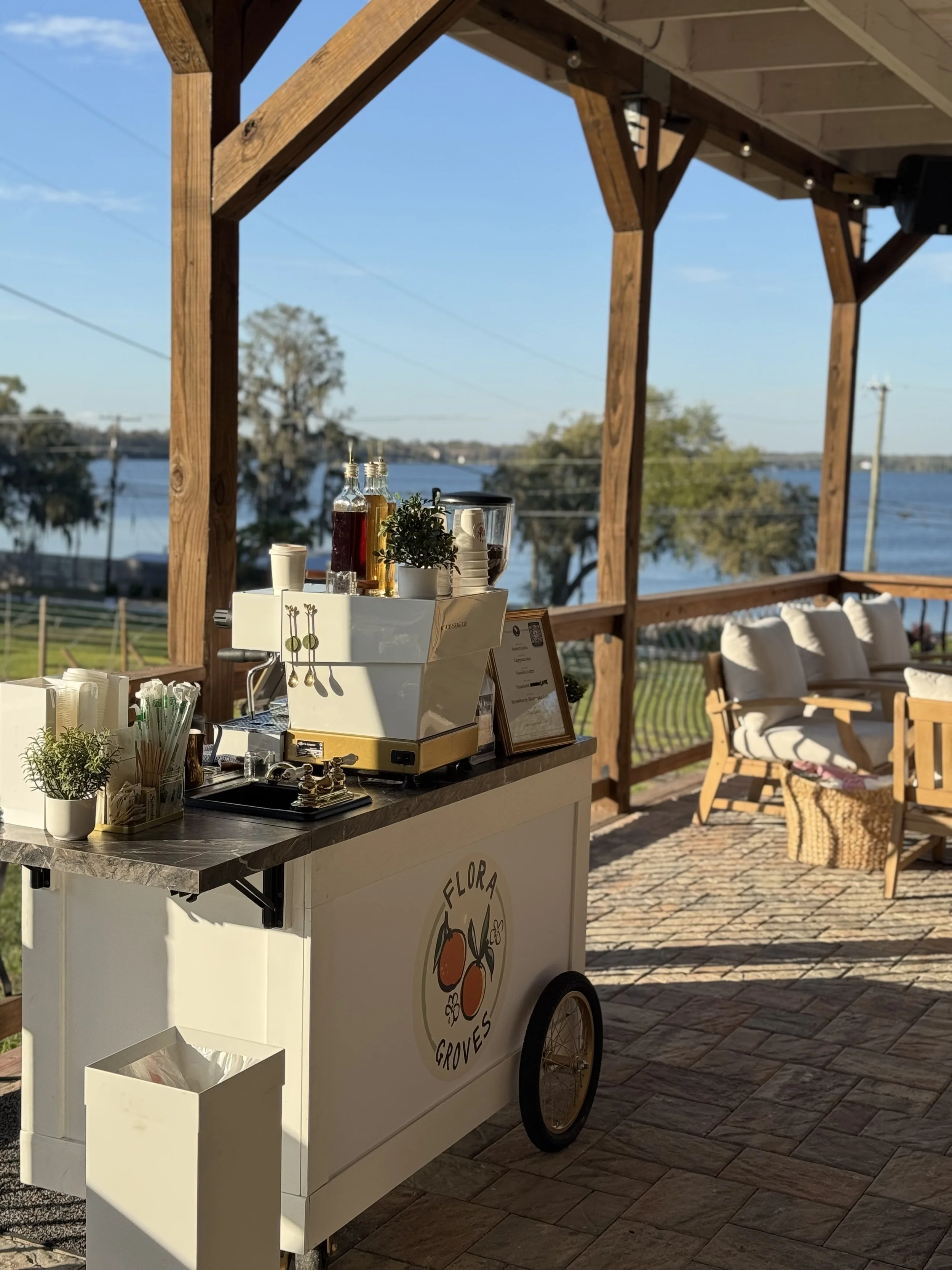 A mobile coffee bar with a logo that reads 'Flora Groves,' set up outside on a patio overlooking a river with trees and blue sky in the background.