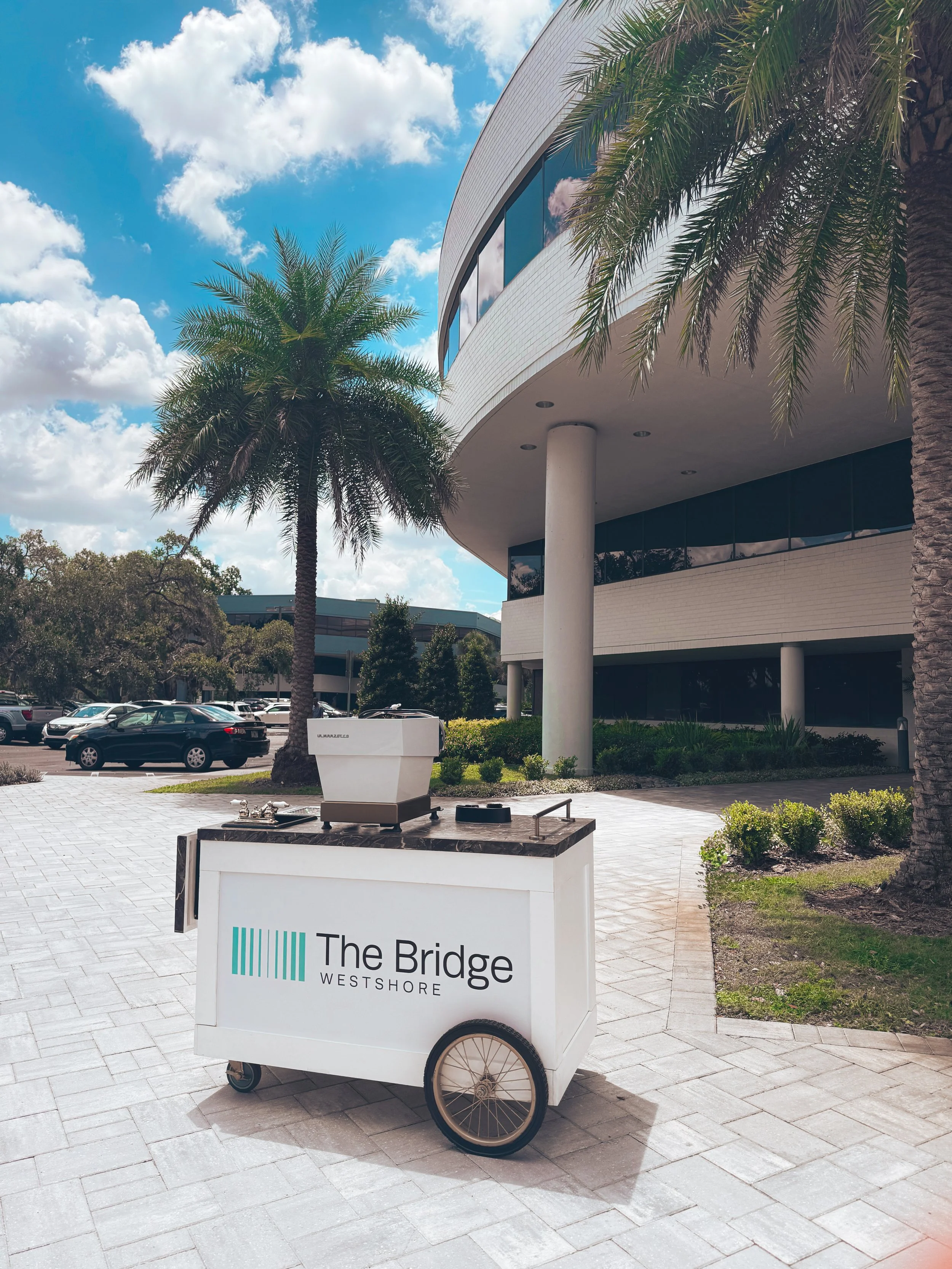 An outdoor scene with a modern white building, palm trees, a parking lot, and a white mobile cart with a sign that reads 'The Bridge Westshore' on a sunny day with blue sky and clouds.