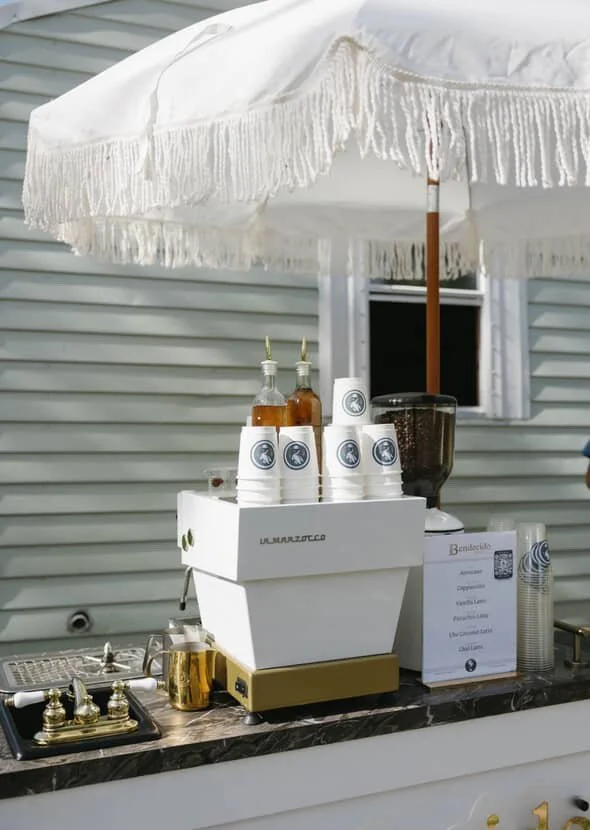 Bar setup with a white umbrella, stacked cups, bottles, and a beverage dispenser on a marble countertop.