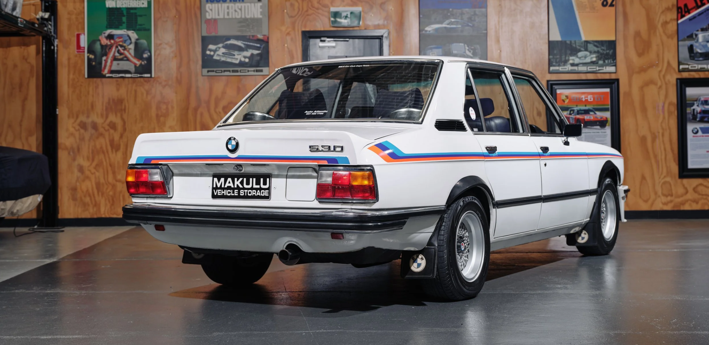 A vintage white BMW 530E car with blue, red, and orange racing stripes, parked indoors against a wooden wall decorated with car posters.