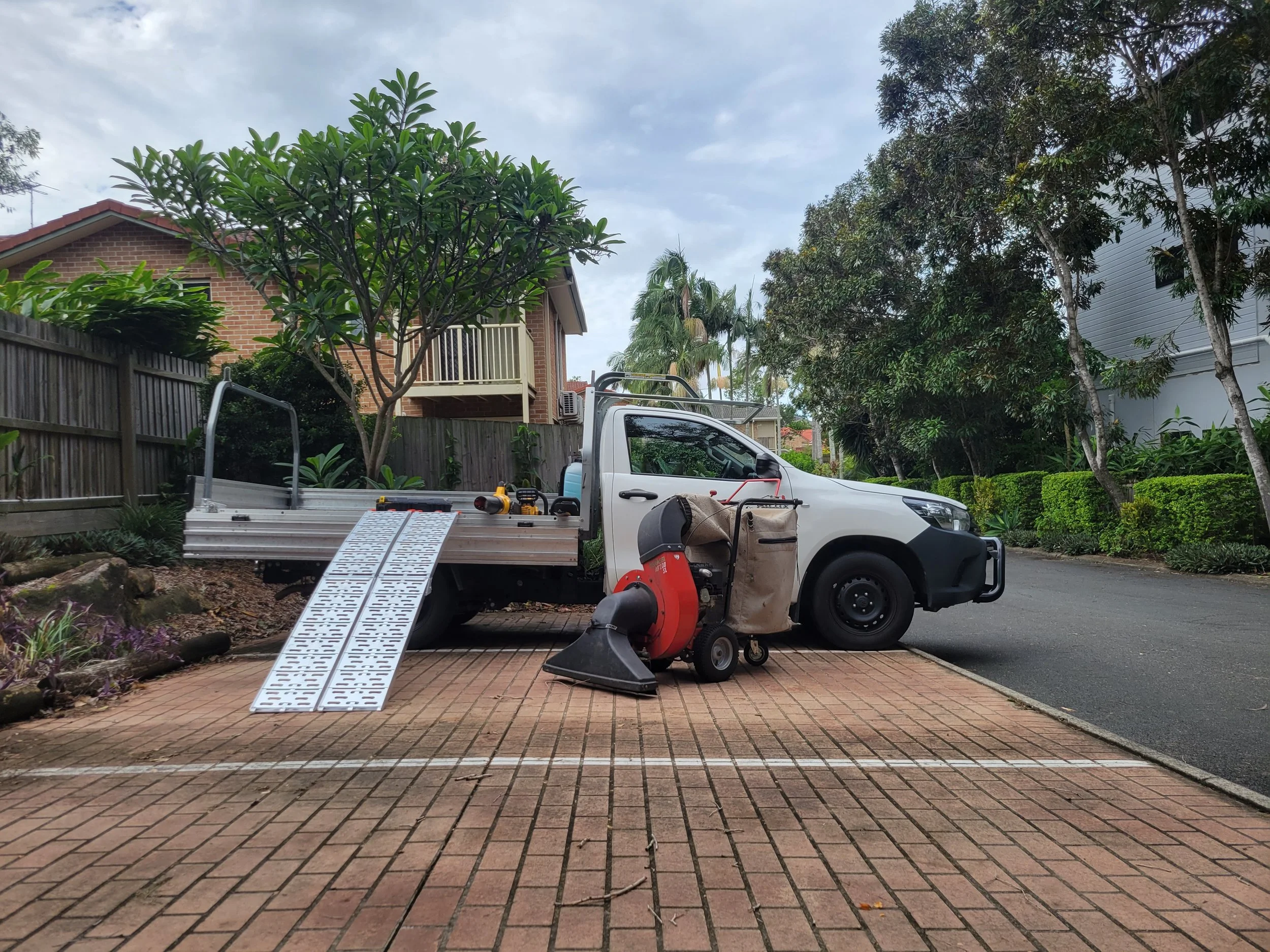 Pickup truck parked on brick driveway, with various tools and equipment for yard work including a leaf blower, leaf vacuum, and a metal ramp. ready for landscape maintenance work