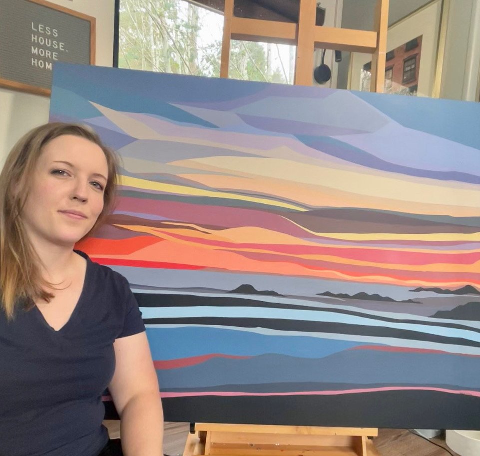 A woman with light brown hair and a black shirt sitting beside a colorful abstract landscape painting featuring a sky with layered clouds in shades of purple, pink, orange, and gray, and a body of water with dark and blue hues, with mountains in the 