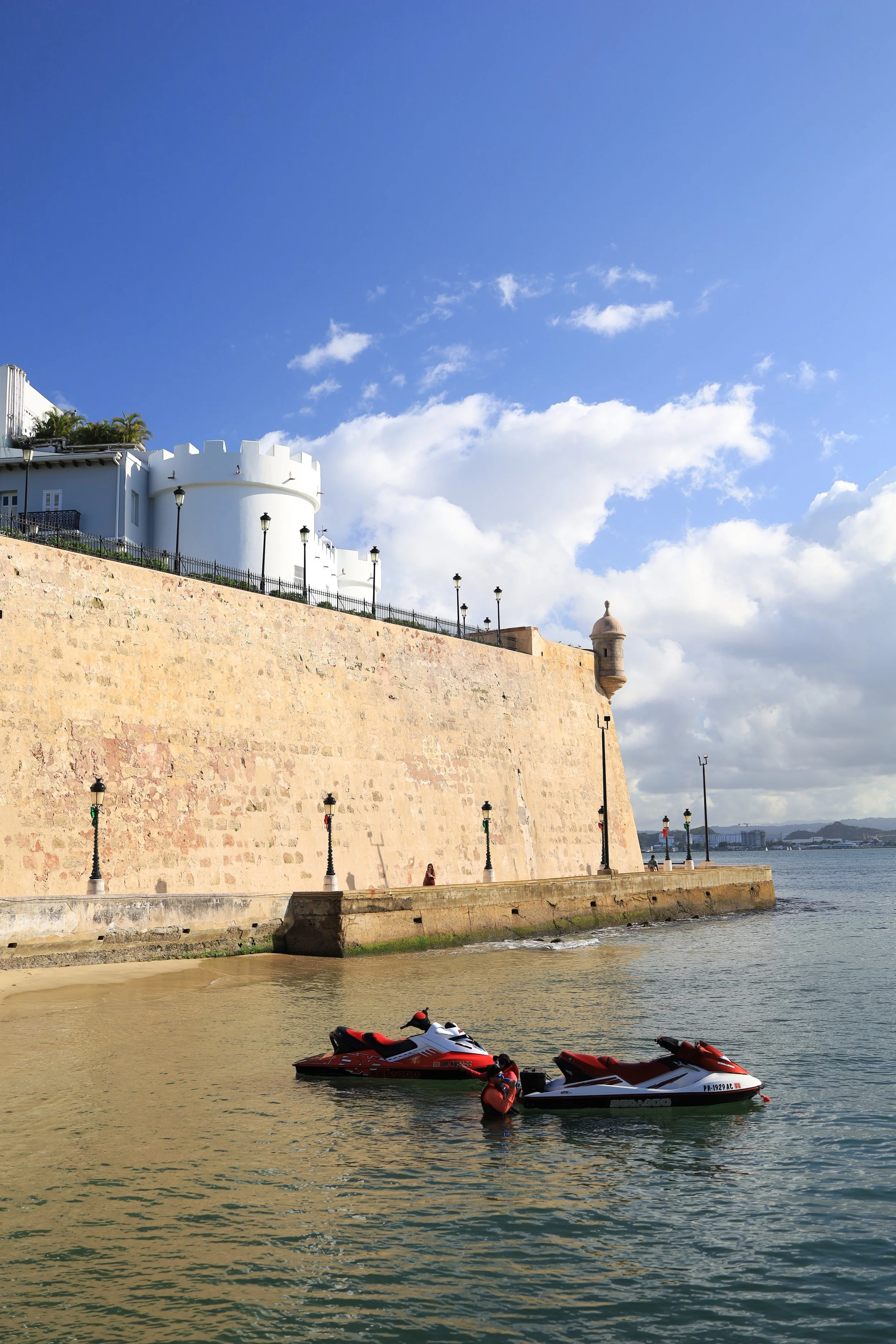 Two jet skis float near the shore of a beach with a high stone wall and a white building with towers in the background under a partly cloudy sky.