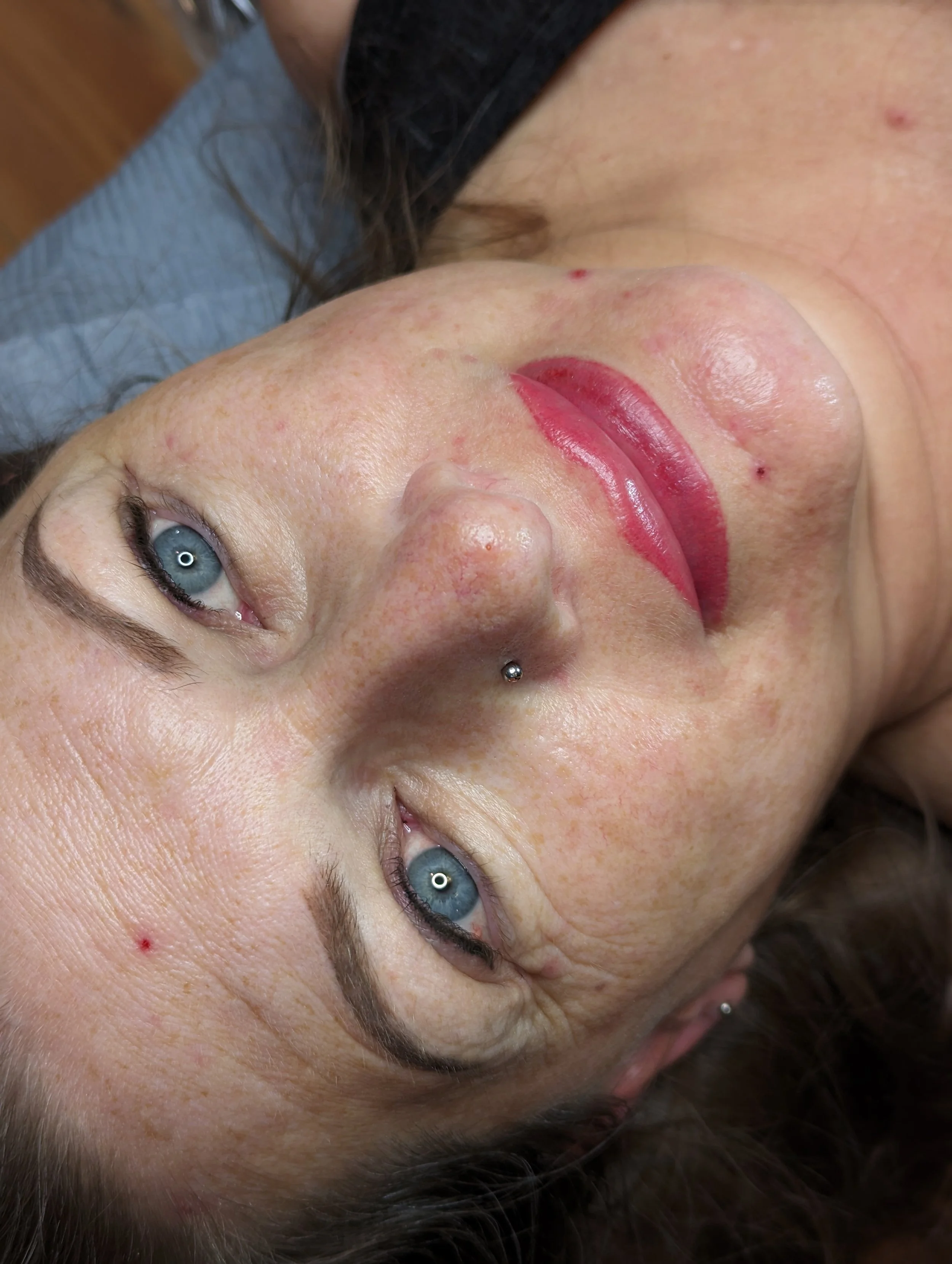 Close-up of a woman lying down with blue eyes, dark eyebrows, light skin, and a septum piercing, showing signs of acne and freckles.