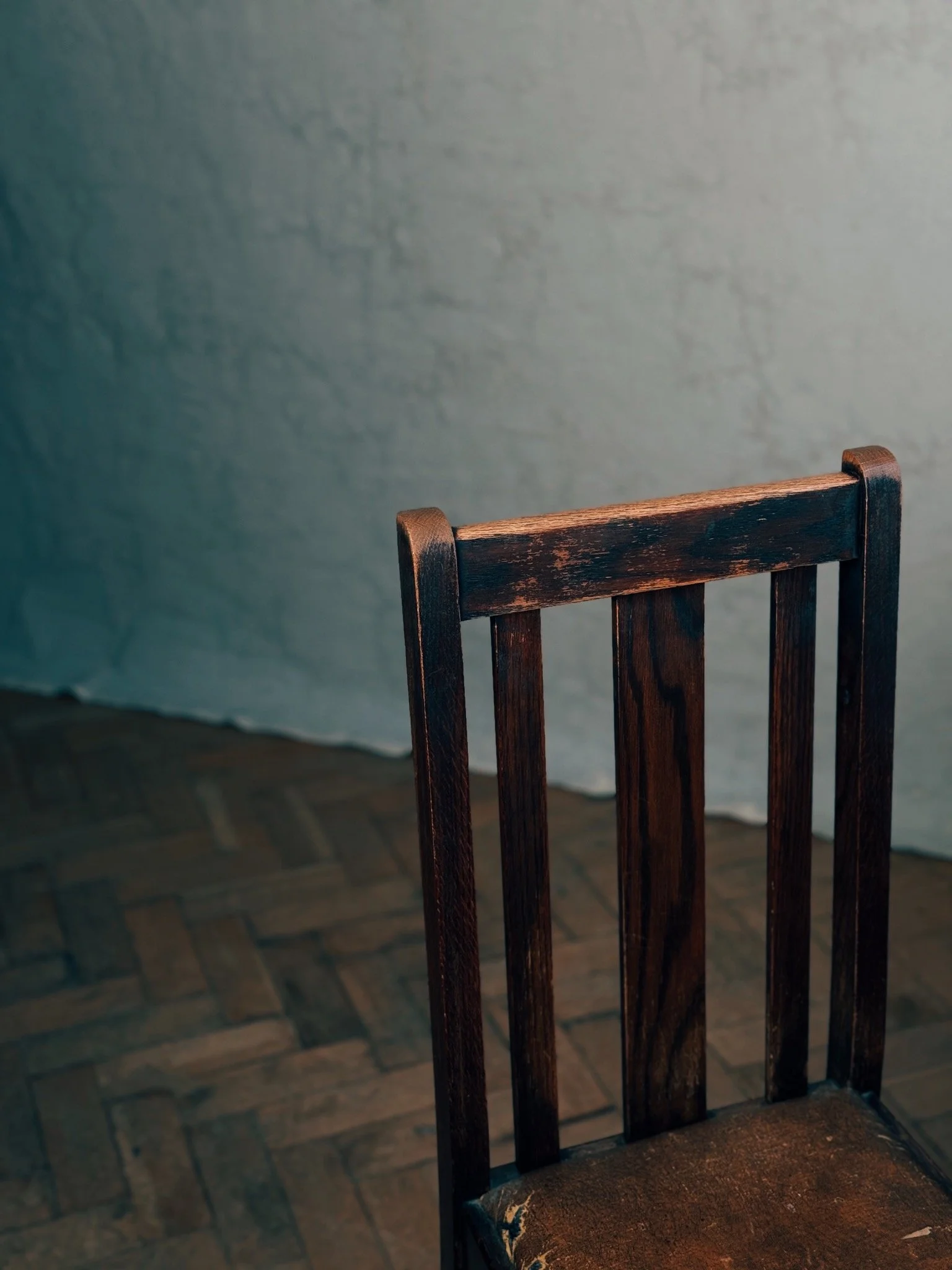 A wooden chair with a worn seat against a plain wall on a wooden floor.