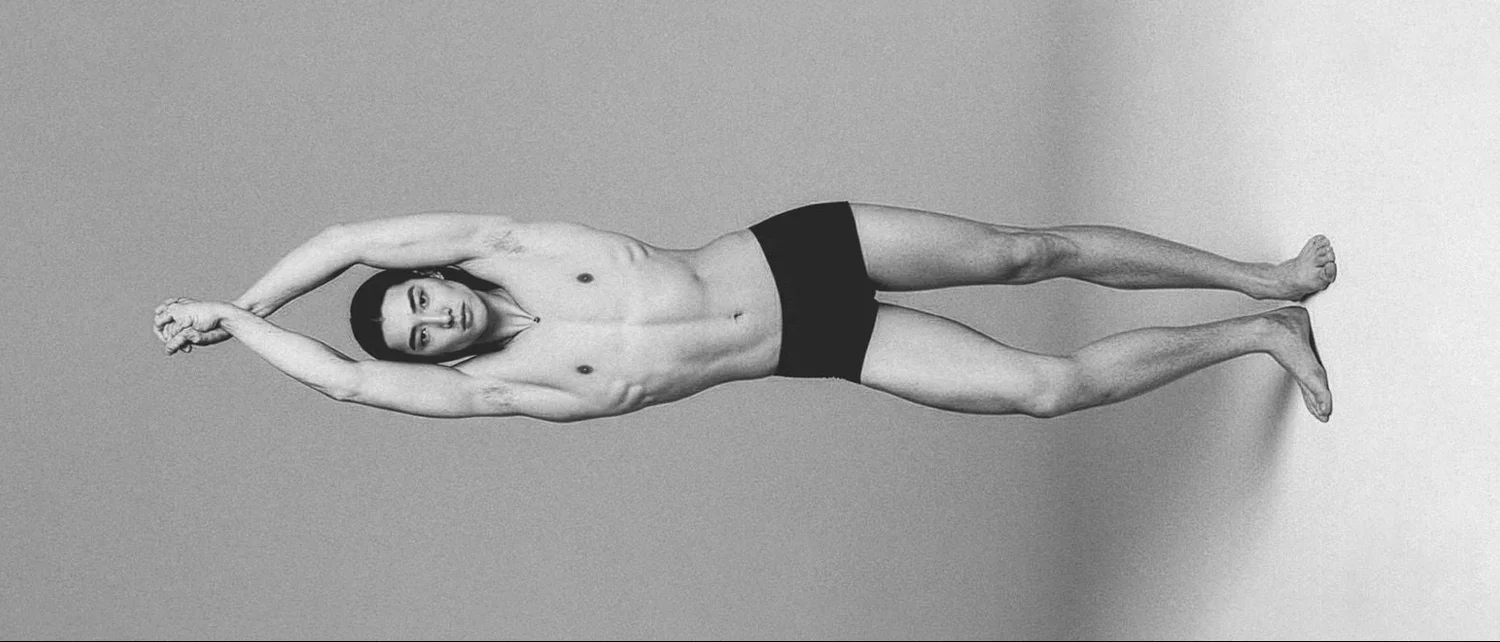 Un homme nu portant uniquement un short noir, allongé sur le côté, avec les bras levés au-dessus de la tête dans une pose de yoga ou de stretching.