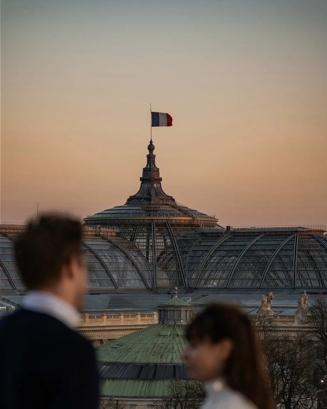 Deux personnes floues, un homme et une femme, devant une architecture en verre d'un bâtiment avec un drapeau français flottant au sommet, sous un ciel au coucher du soleil.