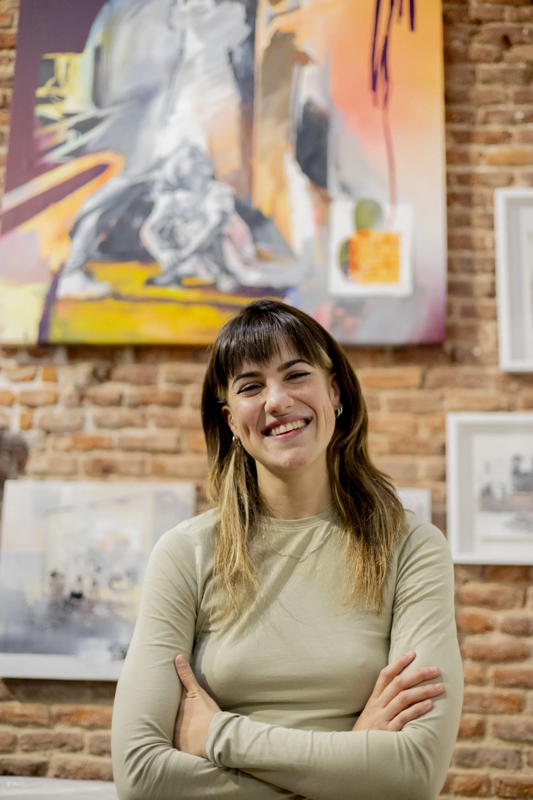 A woman with long brown hair wearing a black shirt and blue gloves looks at an abstract painting in an art studio. The painting features pink, white, orange, and green brush strokes.
