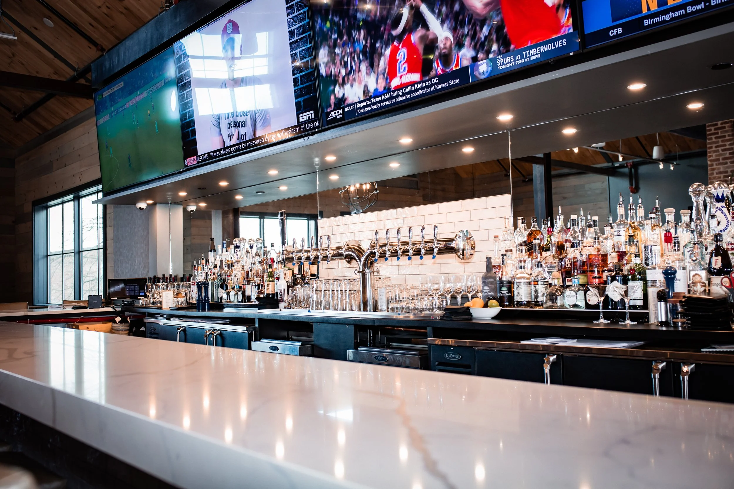 Modern bar interior with liquor bottles, beer taps, and large television screens displaying sports. Elegant countertop with glasses and bar tools.