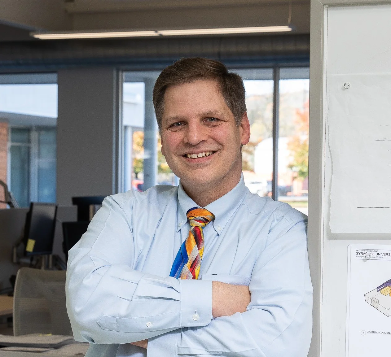 A man with short brown hair, wearing a white dress shirt and a colorful striped tie, smiling in an office environment with cubicles and computers in the background.