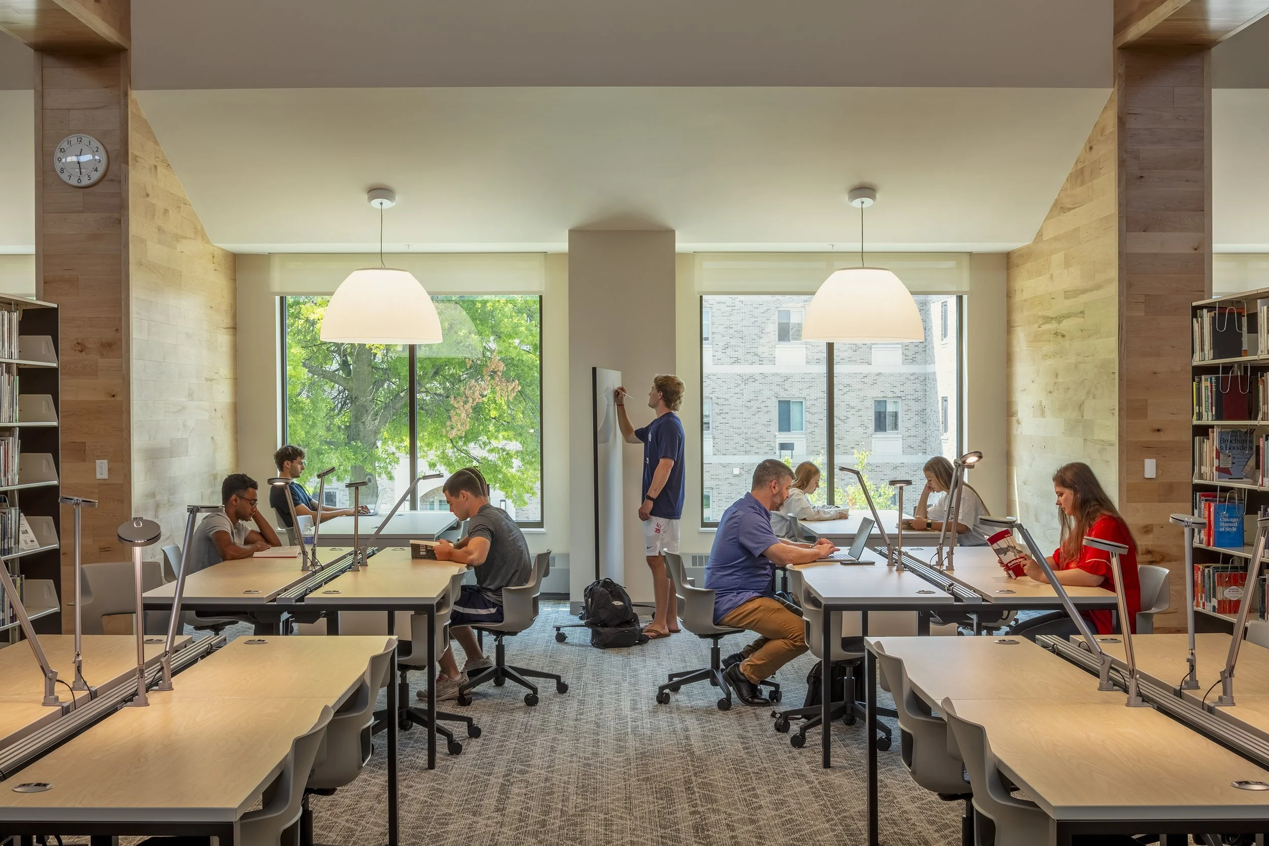 A library study room with eight students working at desks, two standing at a whiteboard, large windows with trees outside, and bookshelves on either side.