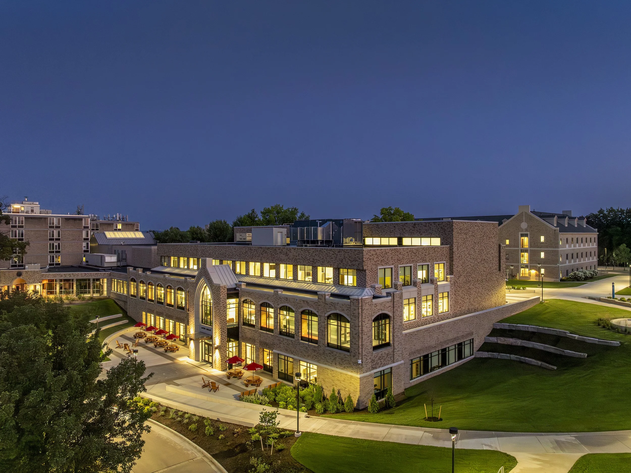 Night view of a multi-story brick building with illuminated windows and outdoor seating area with tables and red umbrellas, surrounded by green lawns and pathways.