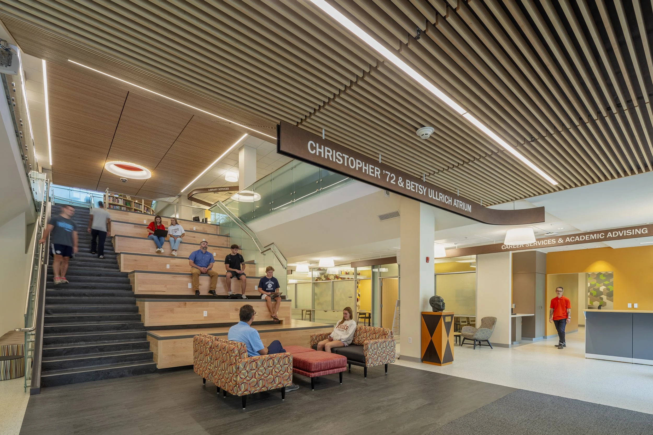 Interior view of a modern educational or institutional building with a wooden slat ceiling, a staircase with wooden seats, and people sitting and walking in a spacious lobby area, including a sitting area with colorful couches and a reception desk.