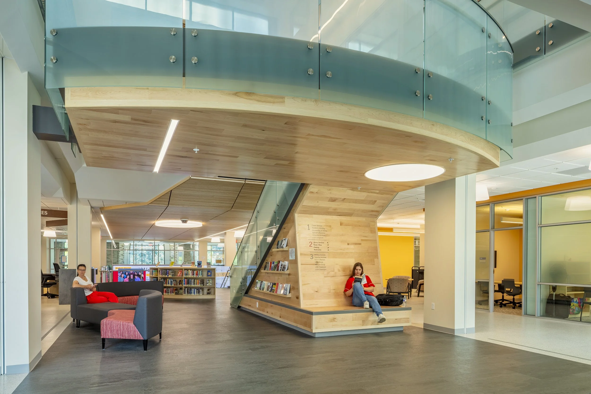 Interior of a modern library or lounge area with a wooden structure, glass railing, seating, and a young woman reading a book.