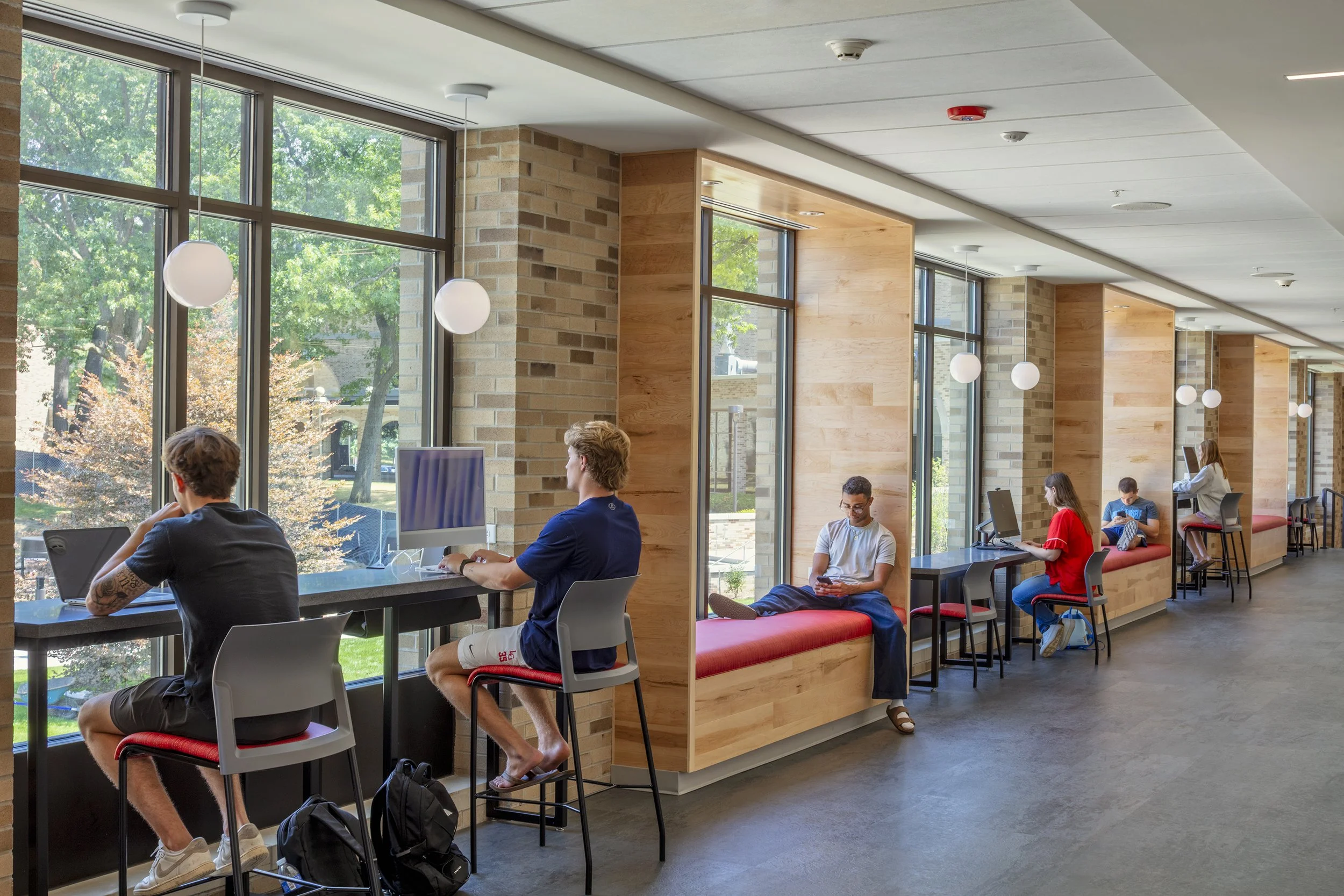 People sitting and working on laptops and smartphones in a bright, modern cafe or waiting area with large windows, brick walls, and wooden accents.