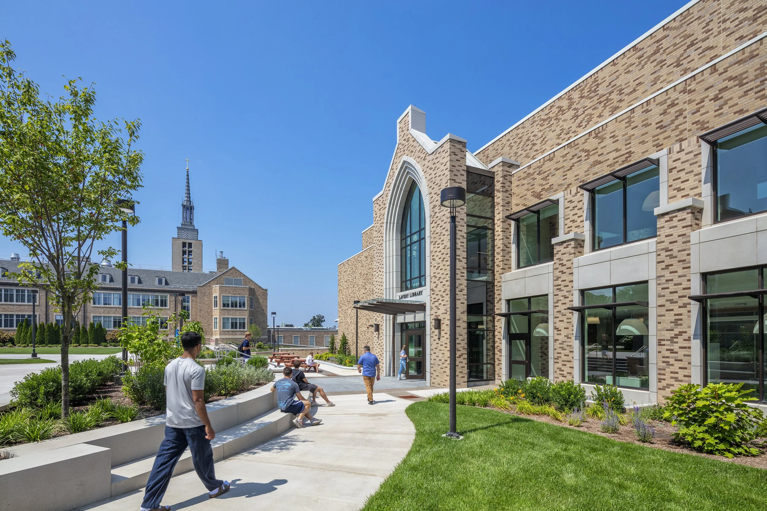 People sitting and walking outside the Liberty Library building, which has large glass windows and a brick facade, with a city skyline and a church spire in the background on a clear sunny day.