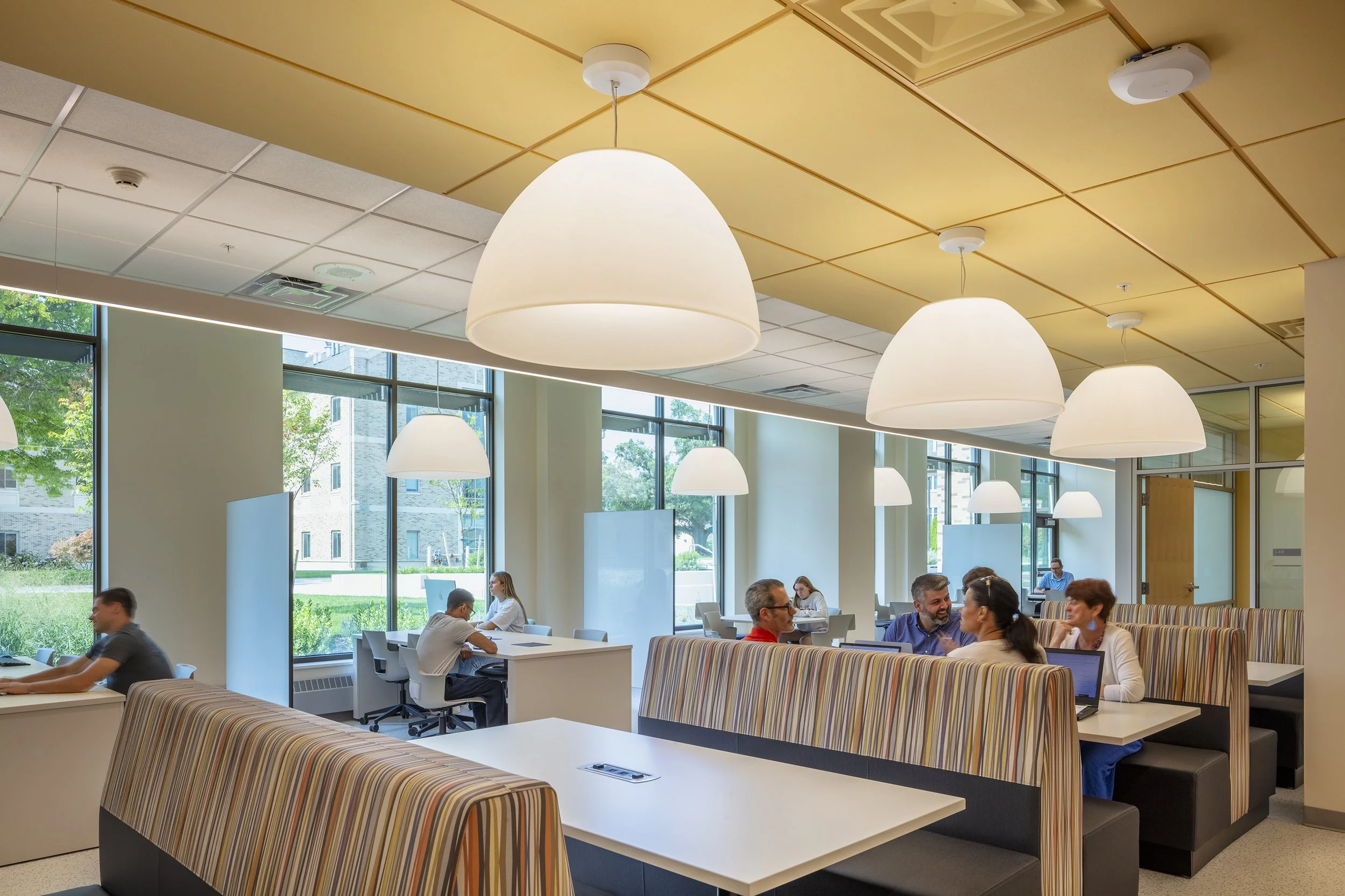 Office cafe or break room with large hanging white lights, ample natural light from large windows, striped booth seating, and people working on laptops and chatting.