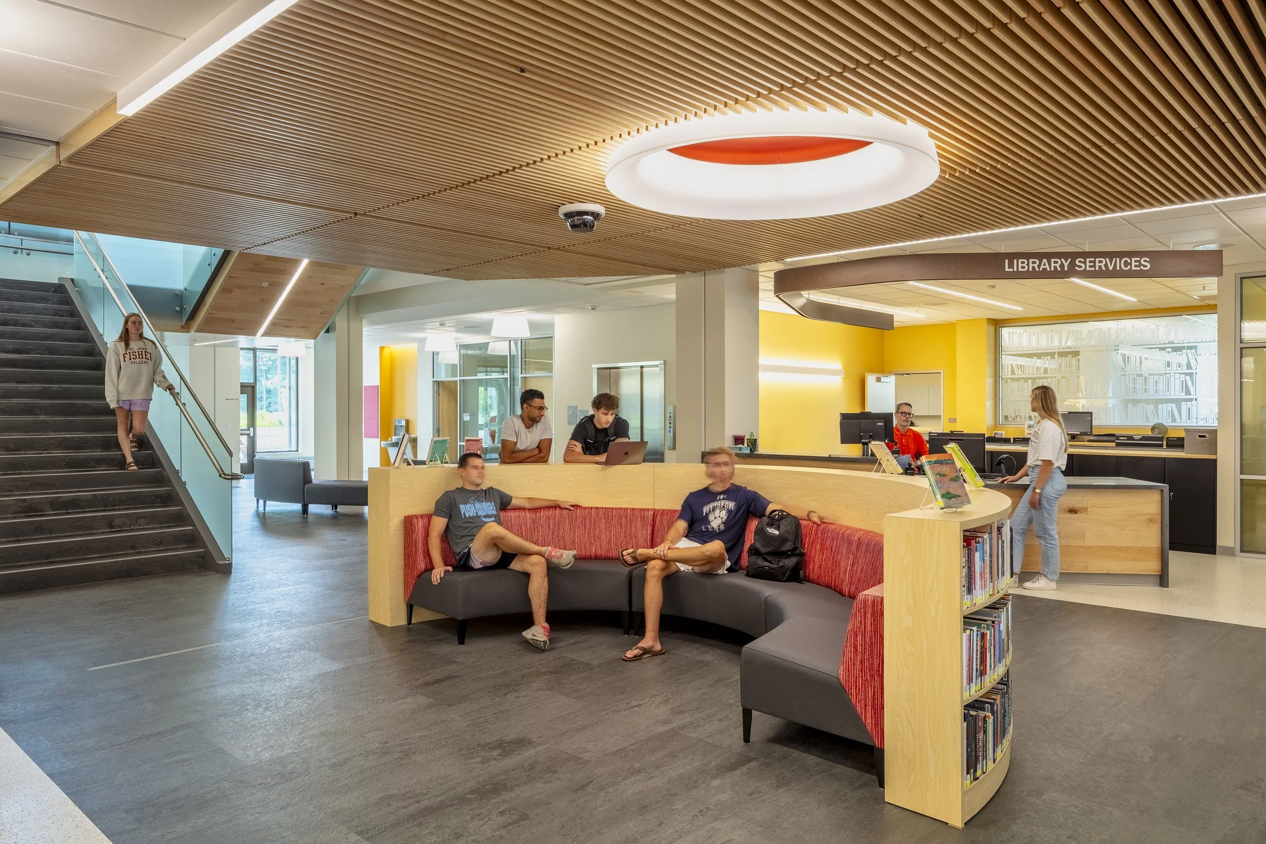 People sitting and standing in a modern library lobby, with a reception desk labeled "Library Services" and a bookshelf.
