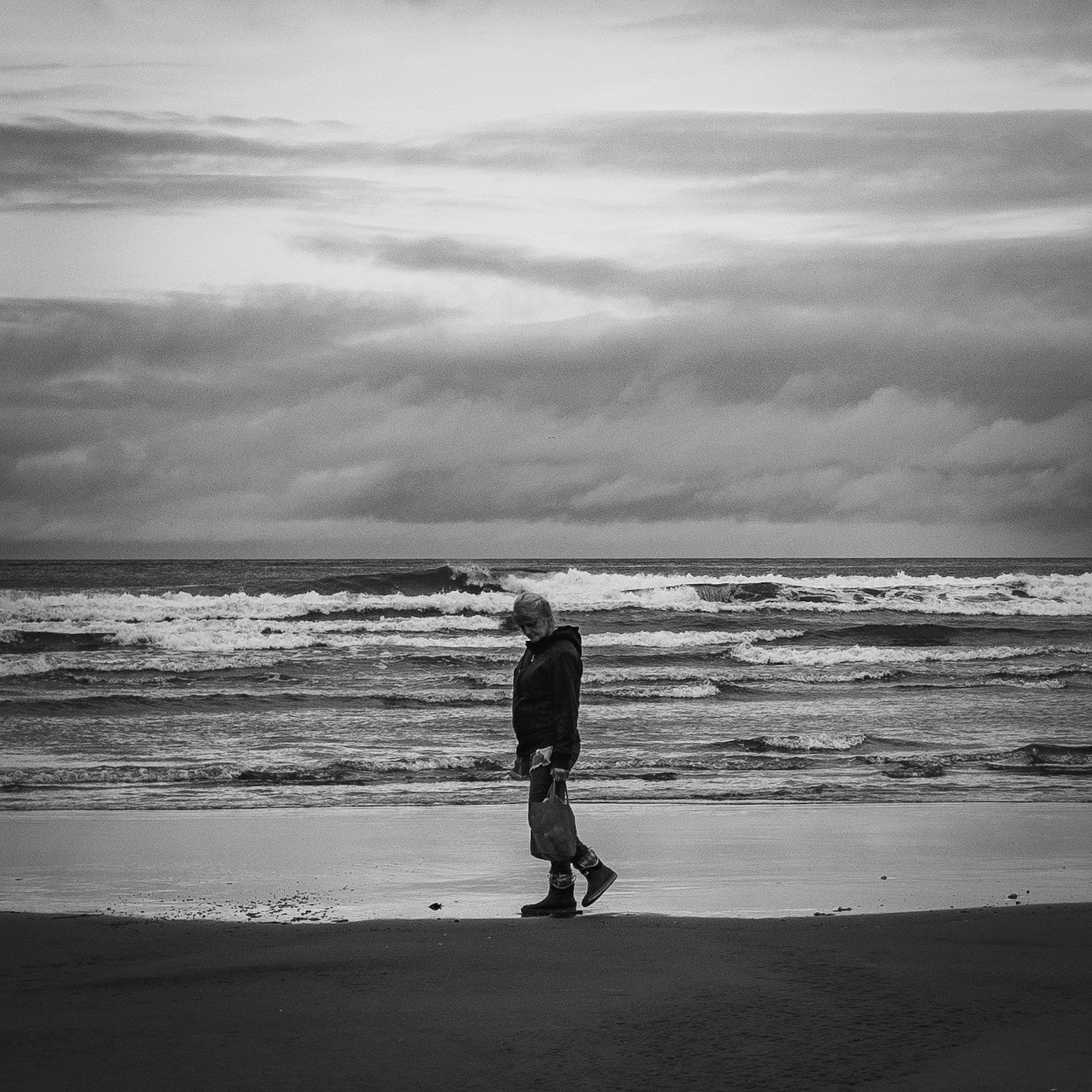 A lone woman cleans the beach of its trash, compiling the wrappers, bottles and other garbage into her bag