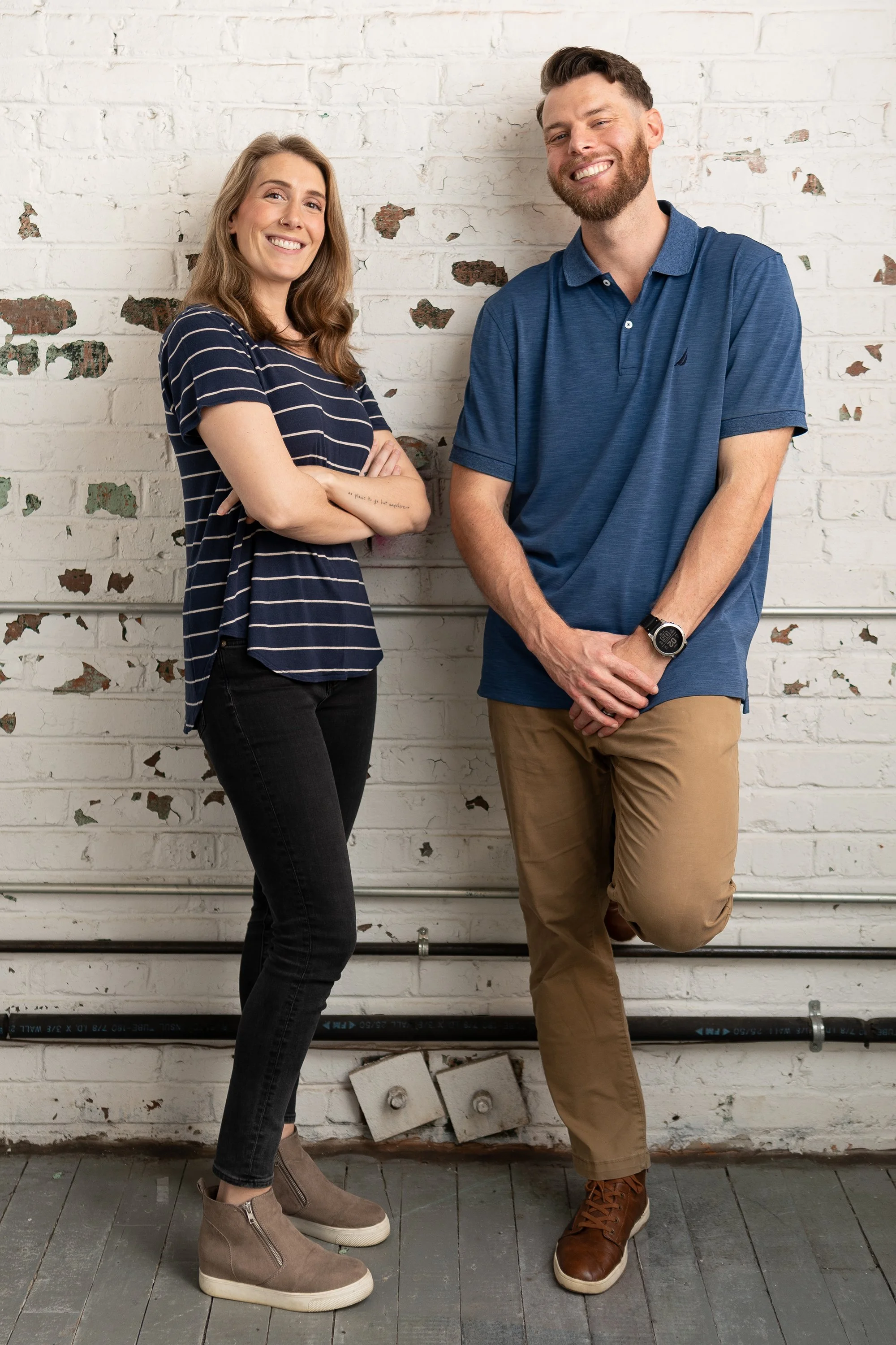 A smiling young woman in a black dress and a young man in a navy suit and sunglasses standing outdoors at sunset.