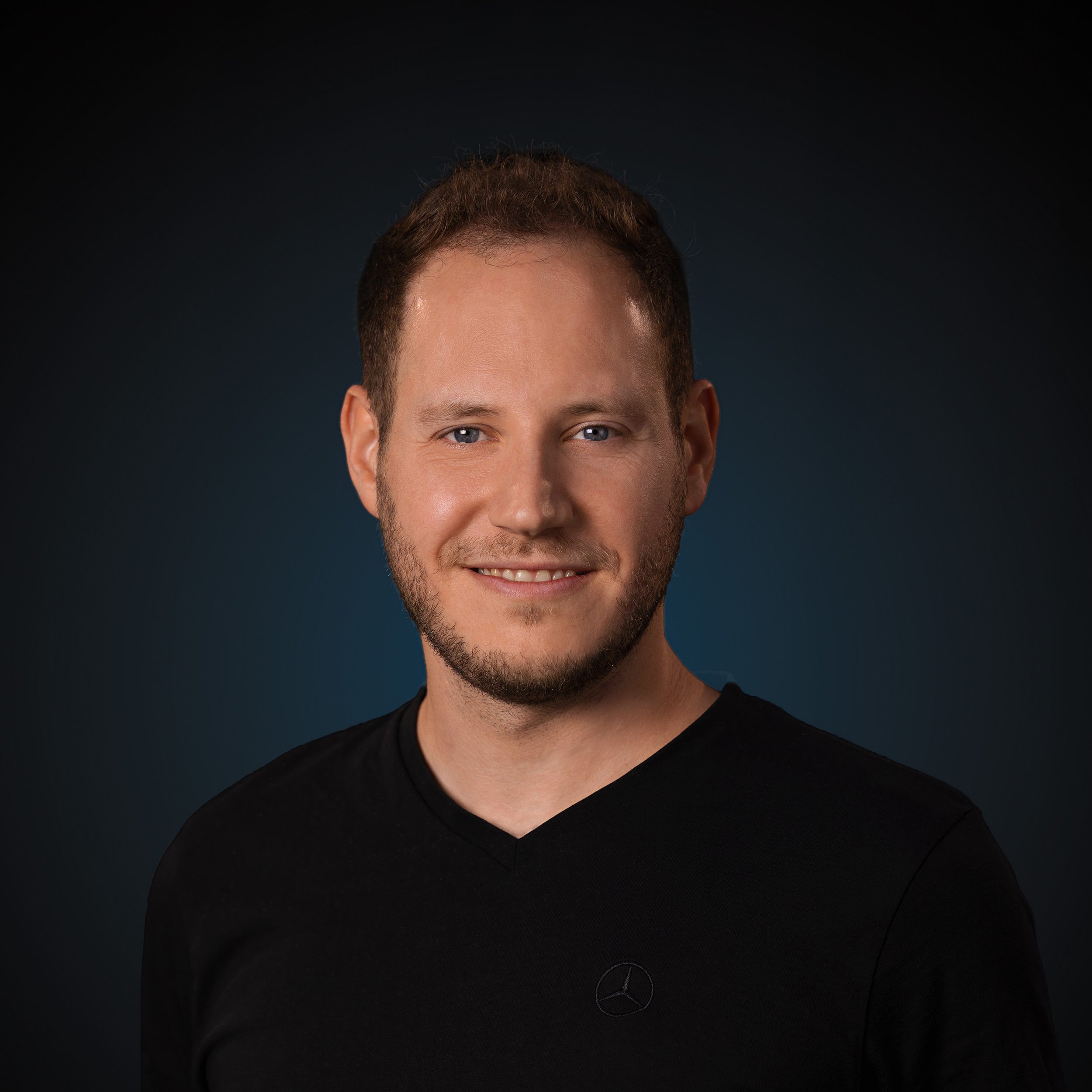 Nahaufnahme des Gesichts eines Mannes mit blauen Augen, dunklem Haar und Bart, trägt einen schwarzen Pullover mit Mercedes-Benz-Logo.
