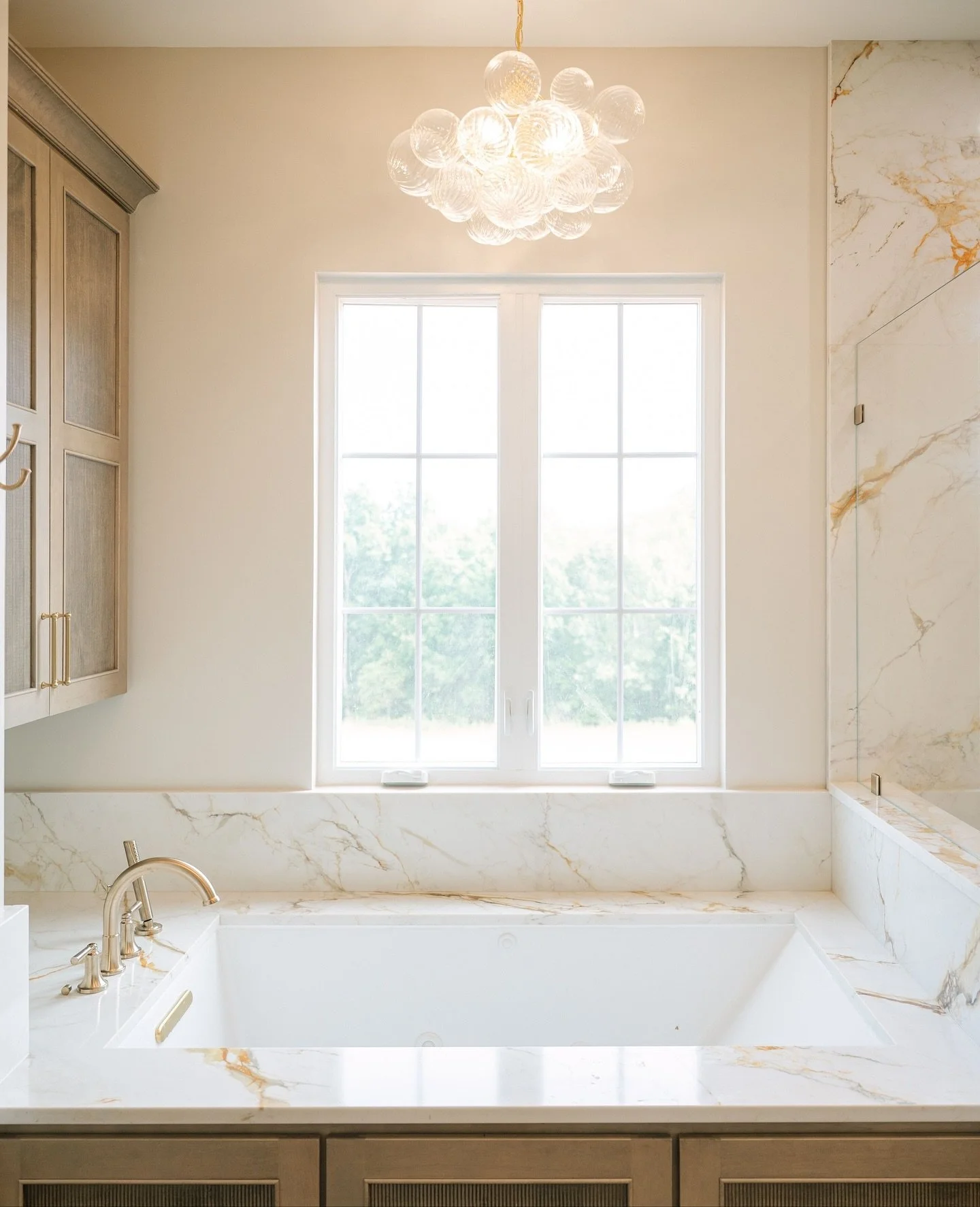 This bathroom is a SHOWSTOPPER. ✨ The kind of space that makes design dreams come true. Macchia Vecchia stone steals the show across the counter tops and full slab shower, while warm cashew-stained cabinetry, champagne bronze hardware, and bold light