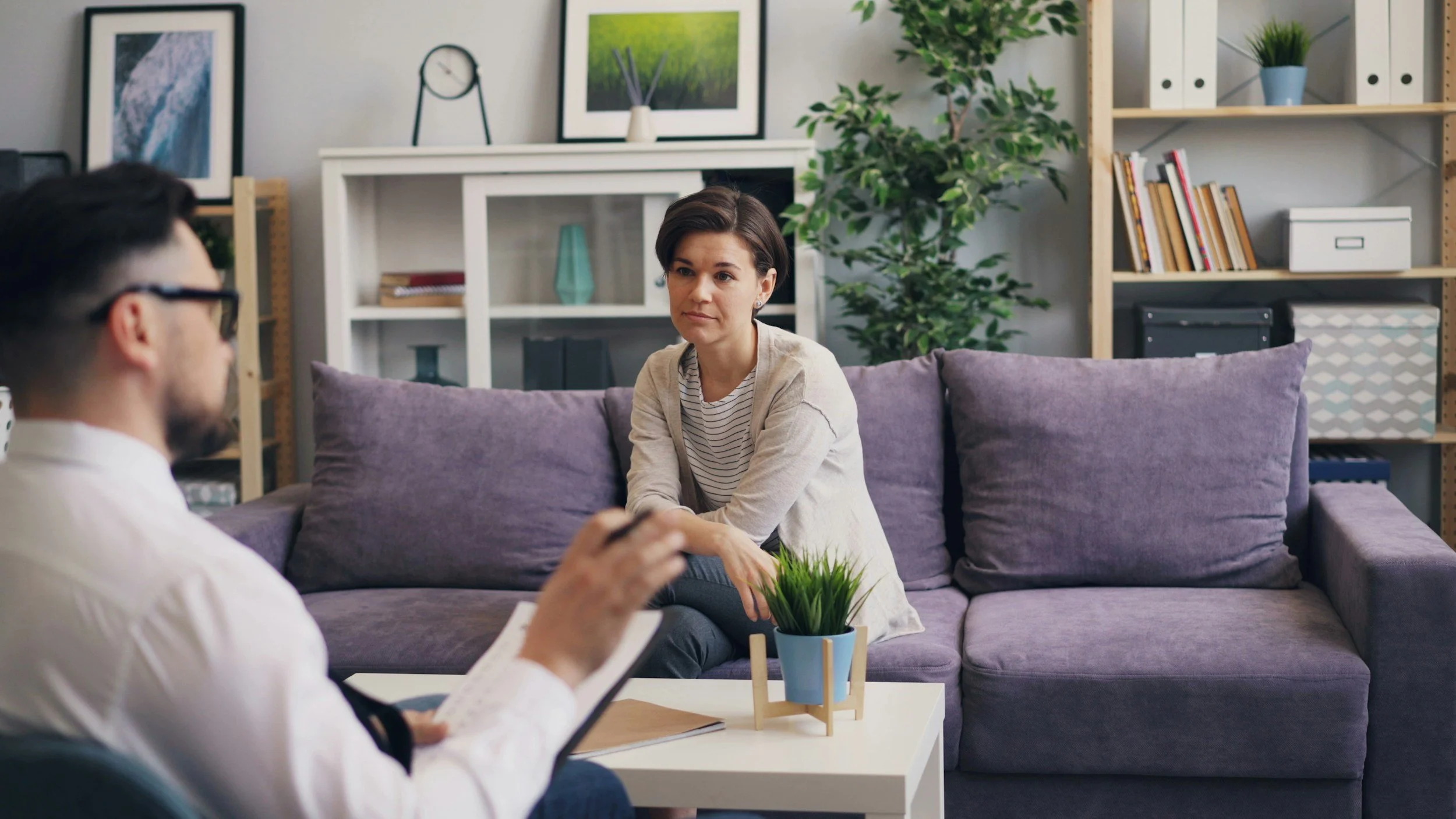 Woman sitting on a gray sofa during a conversation with a man who is taking notes in a notebook, in a cozy living room with decorative pictures and green plants.