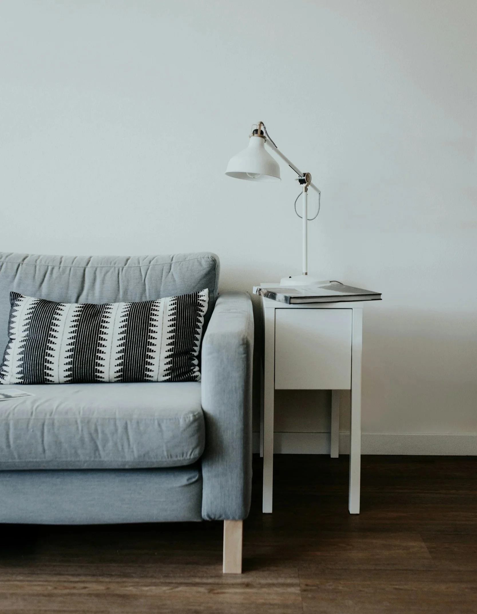 A minimalist bedroom with a white bed, a black and white patterned pillow, a white side table, and a white adjustable desk lamp against a plain white wall and dark hardwood floor.