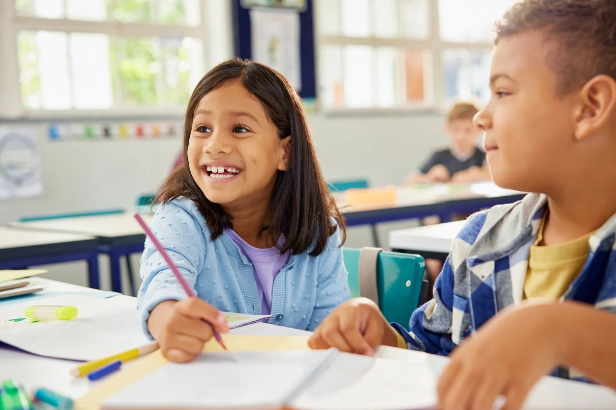 Two children smiling and talking in a classroom, with books and writing supplies on the desks.