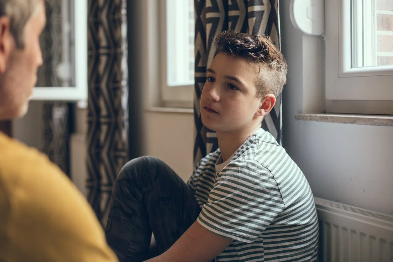 A young boy sitting on the floor by a window, looking thoughtfully at an older person whose face is not visible.