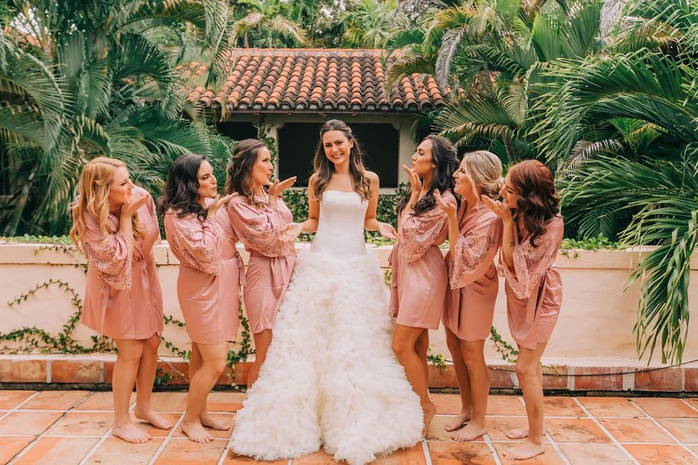 Bride in a white wedding dress standing with six women in pink robes. They are outdoors on a tiled patio with lush green plants and trees in the background.
