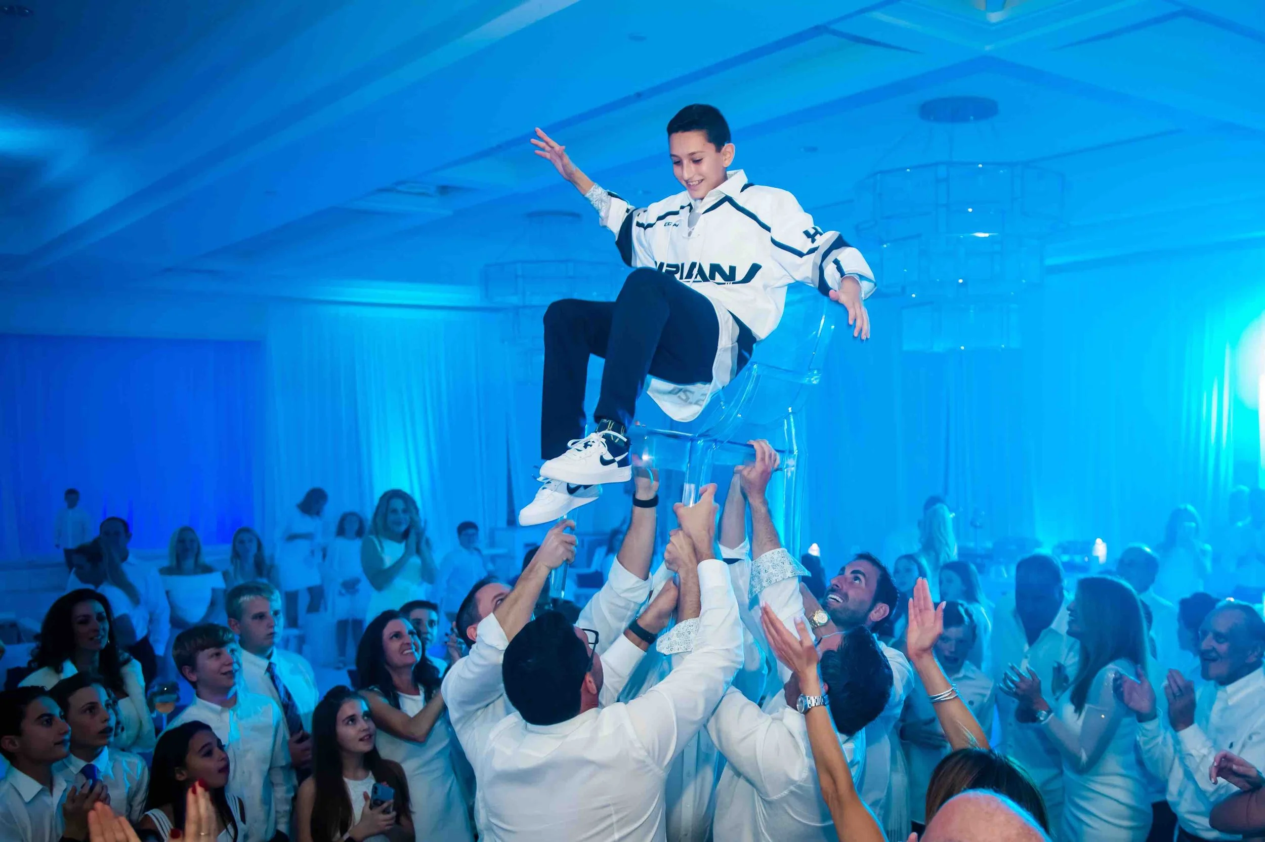 A young boy wearing a white hockey jersey is sitting on a transparent chair balanced on the hands of adults at a party or celebration. The room has blue lighting, and many people are gathered around, clapping and watching as he is lifted.
