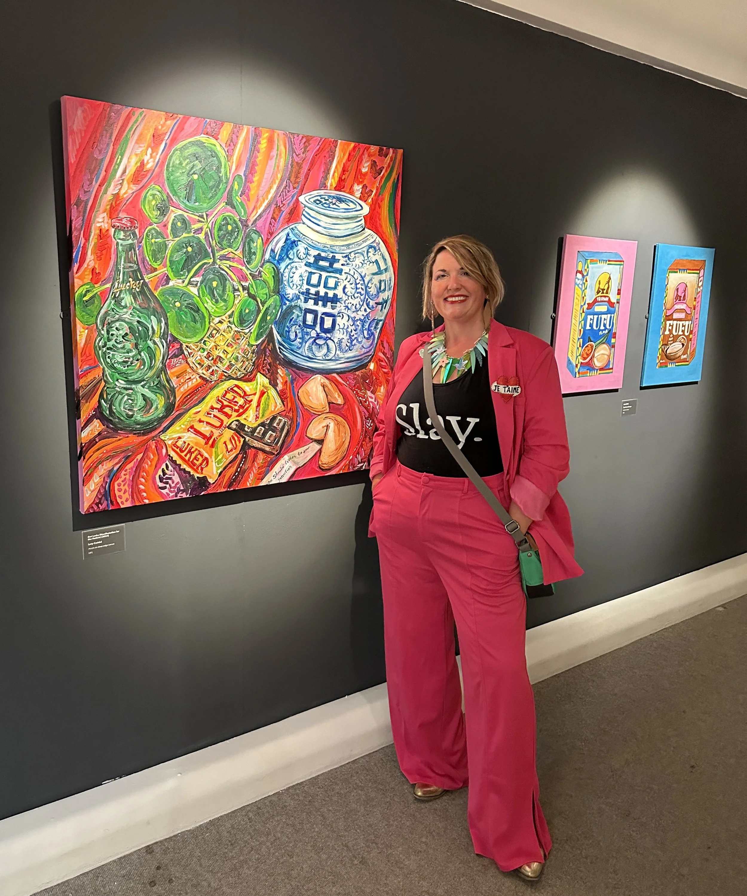 A woman in a bright pink suit stands smiling in an art gallery, with colorful still life paintings behind her. The prominent painting features green grapes, a blue and white jar, a green bottle, and various snacks, painted with vivid, energetic brushstrokes.