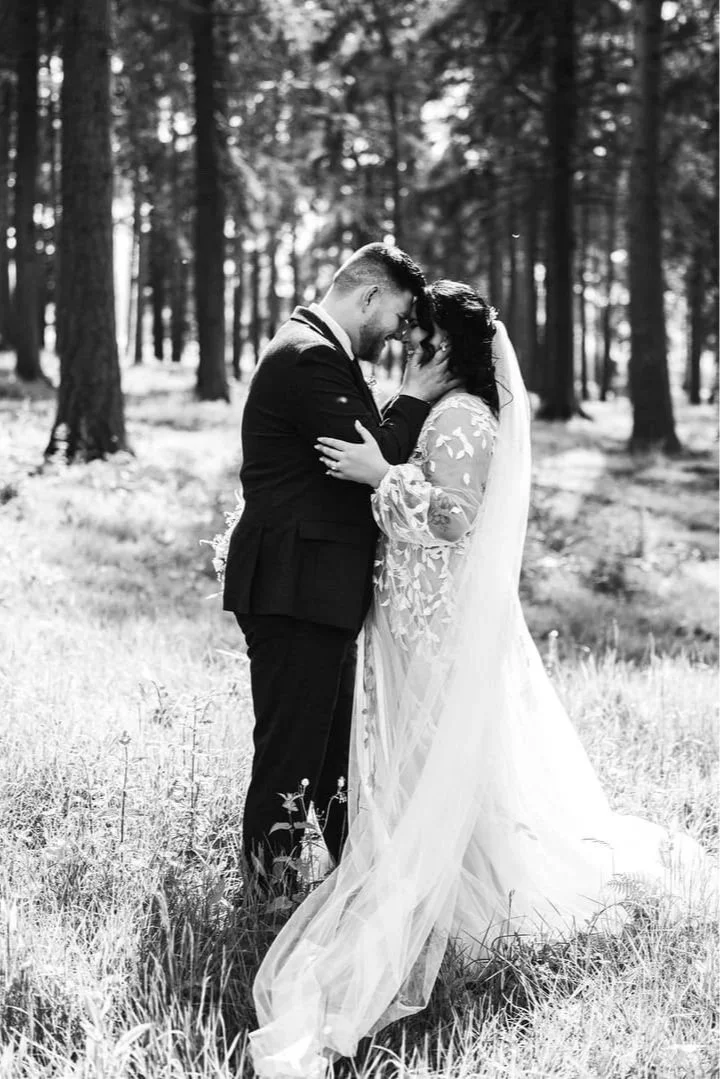 A couple in wedding attire sharing a kiss and touching foreheads in a forested area with sunlight filtering through the trees.