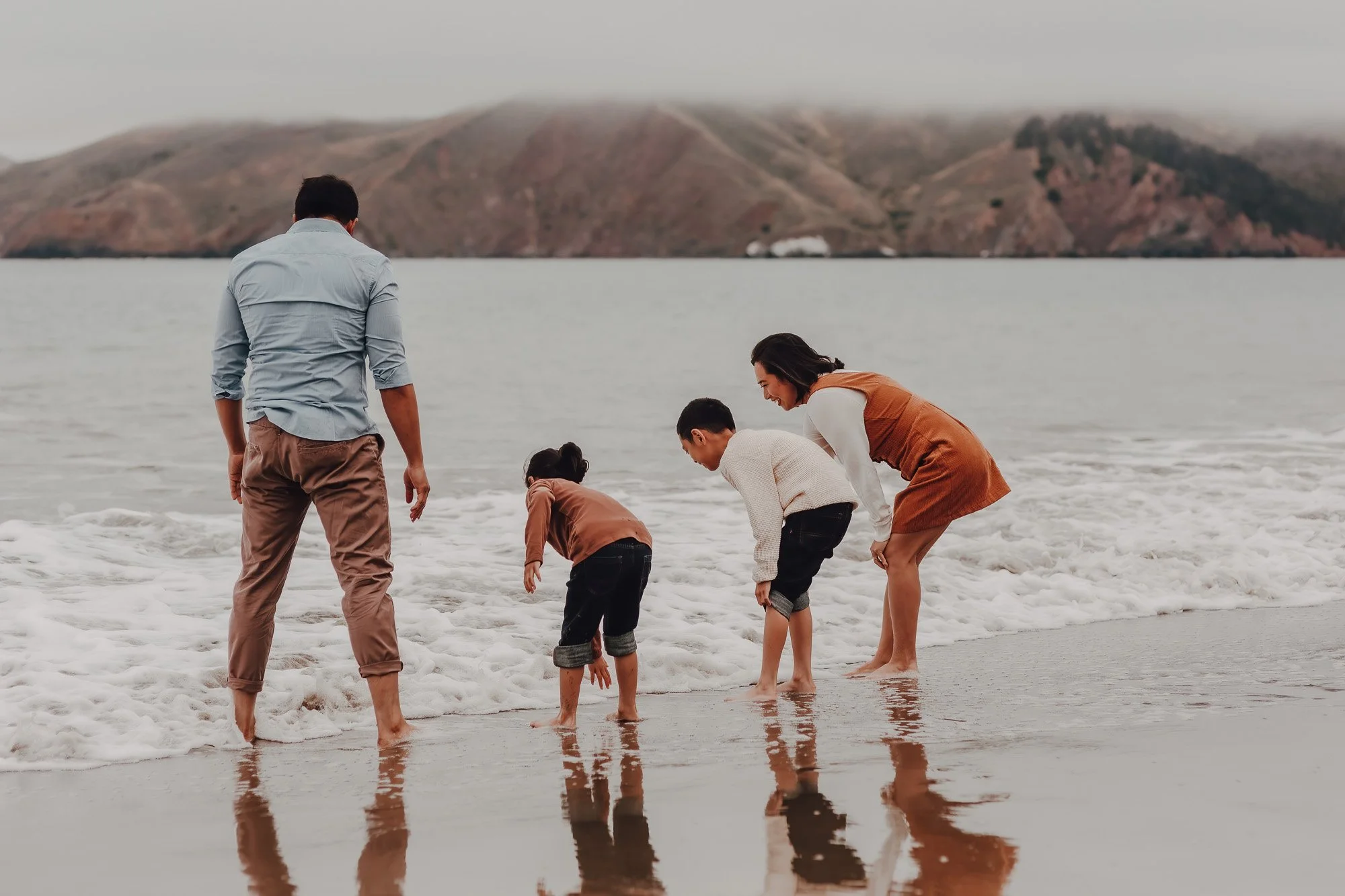 Family of four playing at the beach, with two children bending over near the water and two adults standing nearby, with mountains in the background.