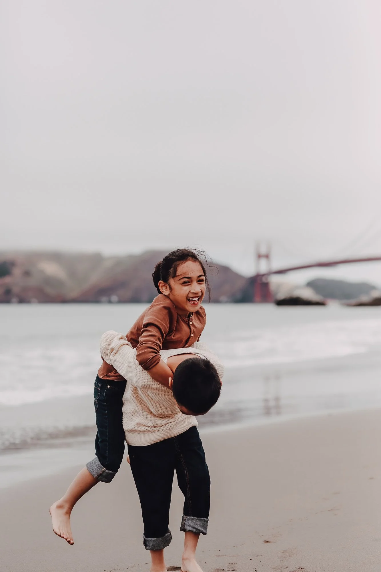 A girl and a boy playing on the beach with the Golden Gate Bridge in the background. The girl is riding on the boy's back, both are smiling and having fun.
