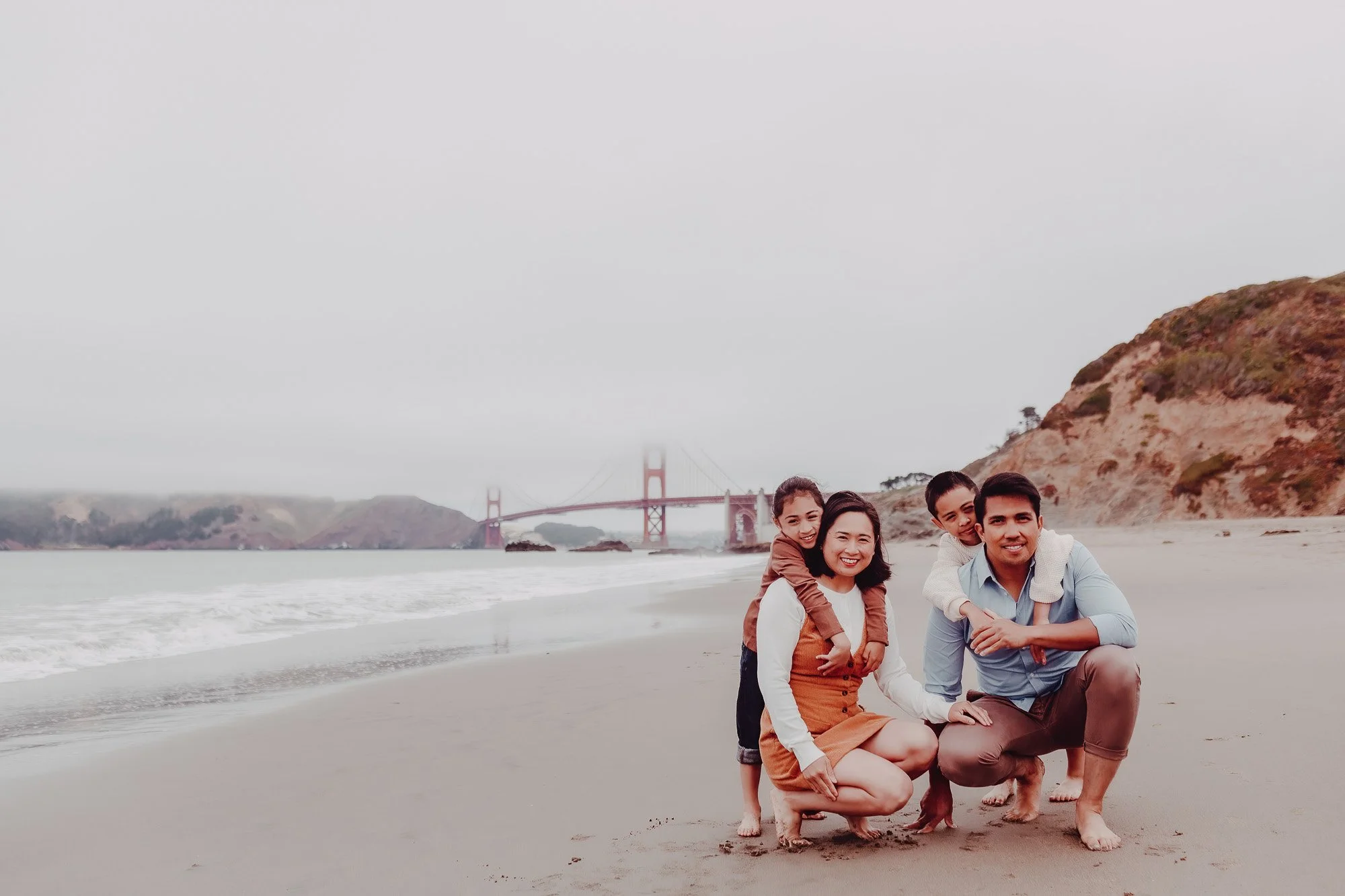 A family of four posing on a beach with the Golden Gate Bridge in the background, smiling and dressed casually.