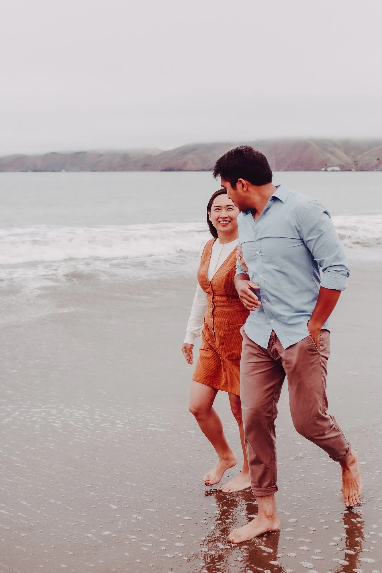 A smiling woman and a man walking barefoot on the beach near the water, with hills visible in the background.