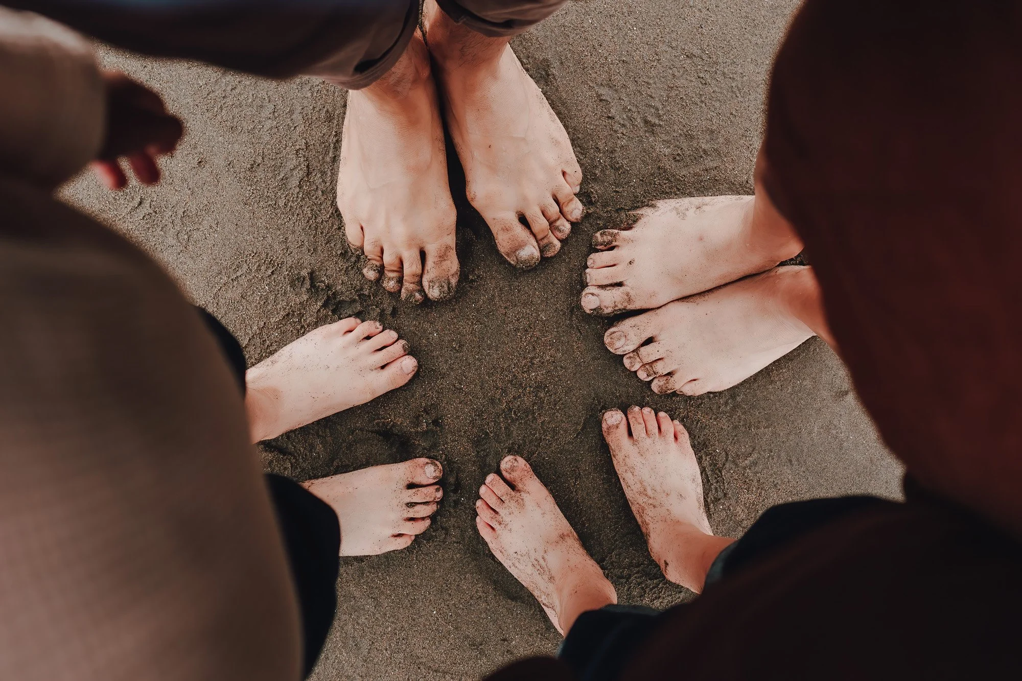 Six people standing in a circle with their bare feet on sandy ground, some feet are muddy.